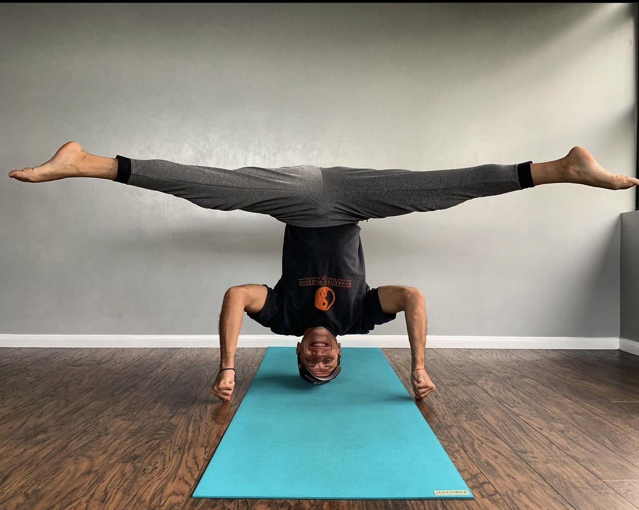 Person balancing on hands with legs extended horizontally, head near blue yoga mat, in a room with plain wall.