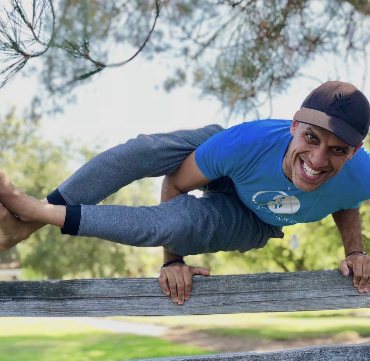 Person balancing on a wooden fence outdoors, smiling, wearing a cap and blue shirt, with trees and sky in background.
