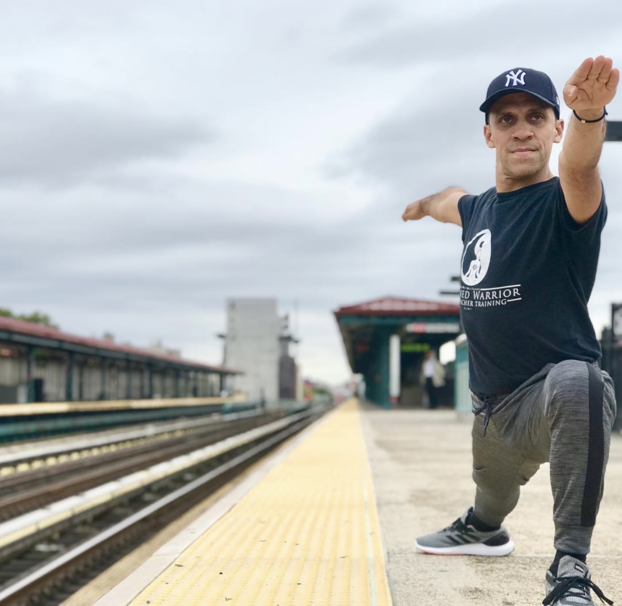 Person in a black t-shirt and gray pants performs a yoga pose on a train platform, with train tracks and a cloudy sky in background.