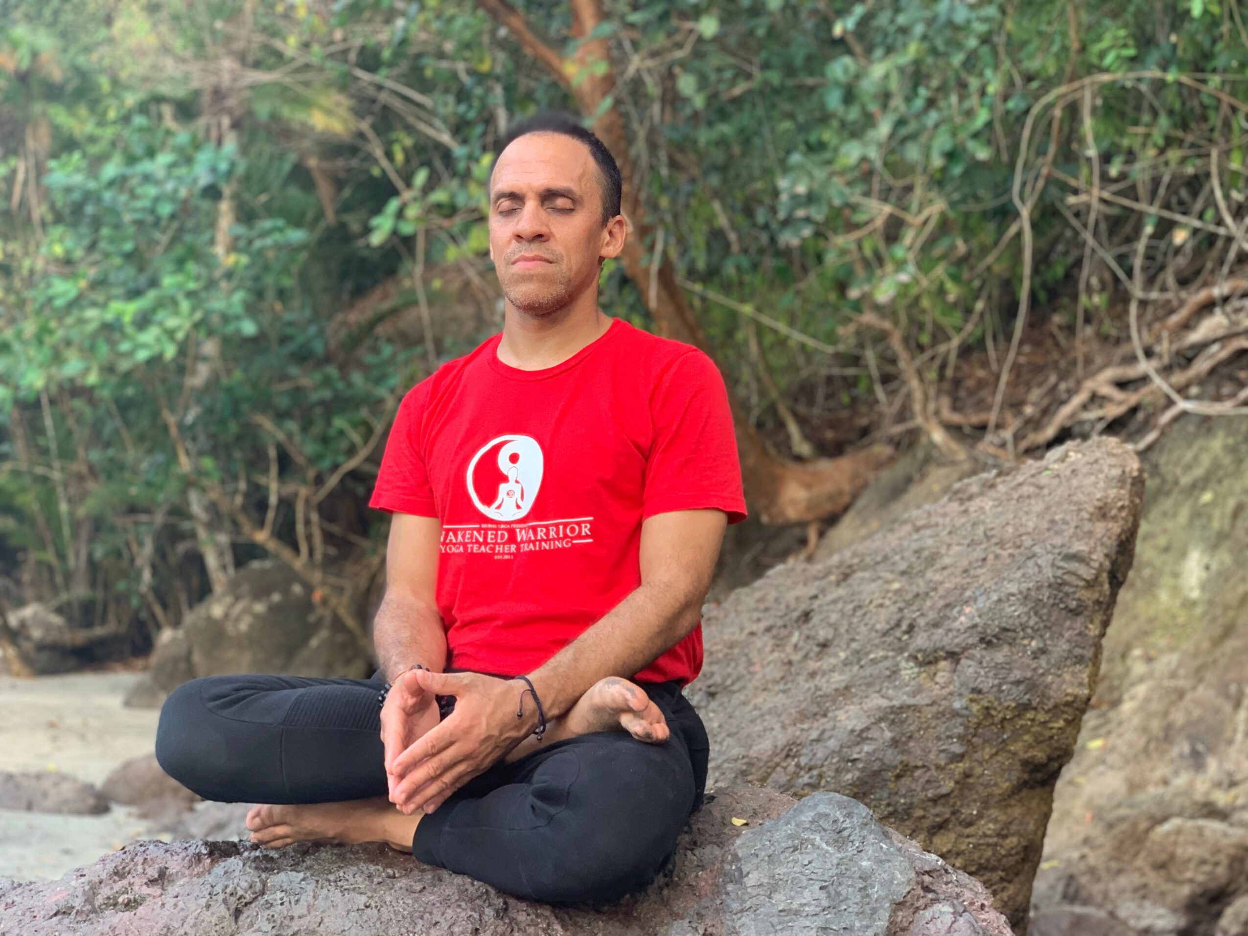 Person sitting cross-legged outdoors with trees and rocks in background, wearing a red T-shirt and black pants.