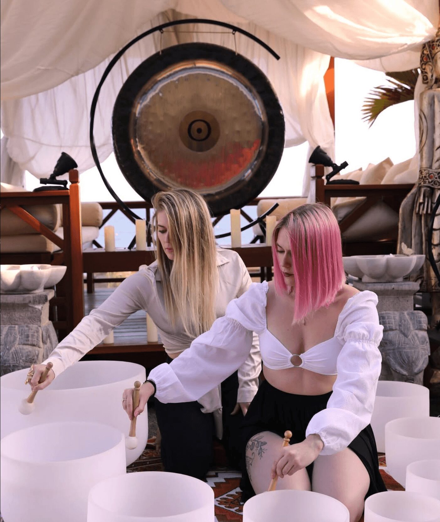 Two women with colorful hair sit among white bowls in a decorated room with a large gong in the background.