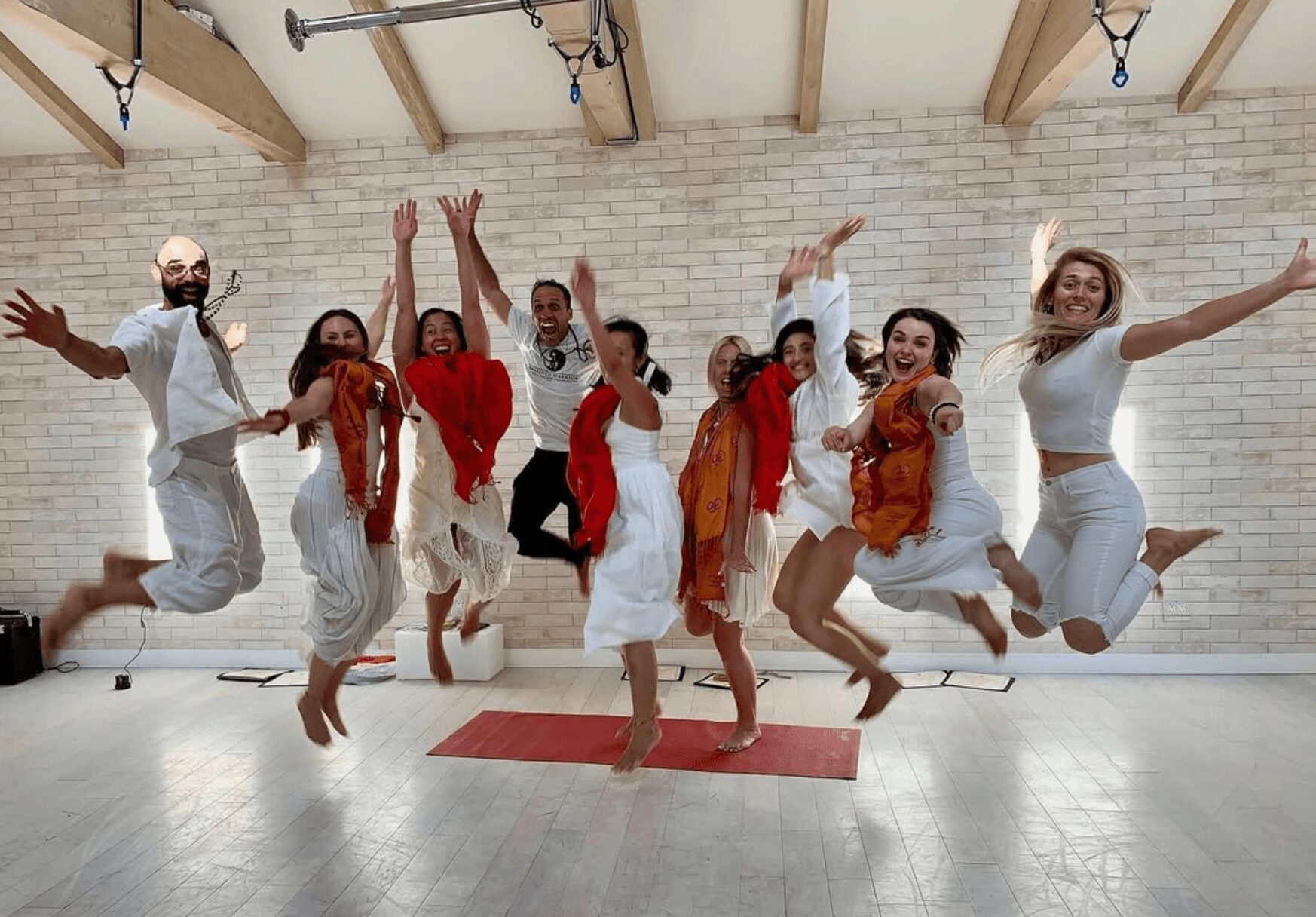 Group of nine people jumping in a dance studio with a brick wall background and wooden ceiling beams.