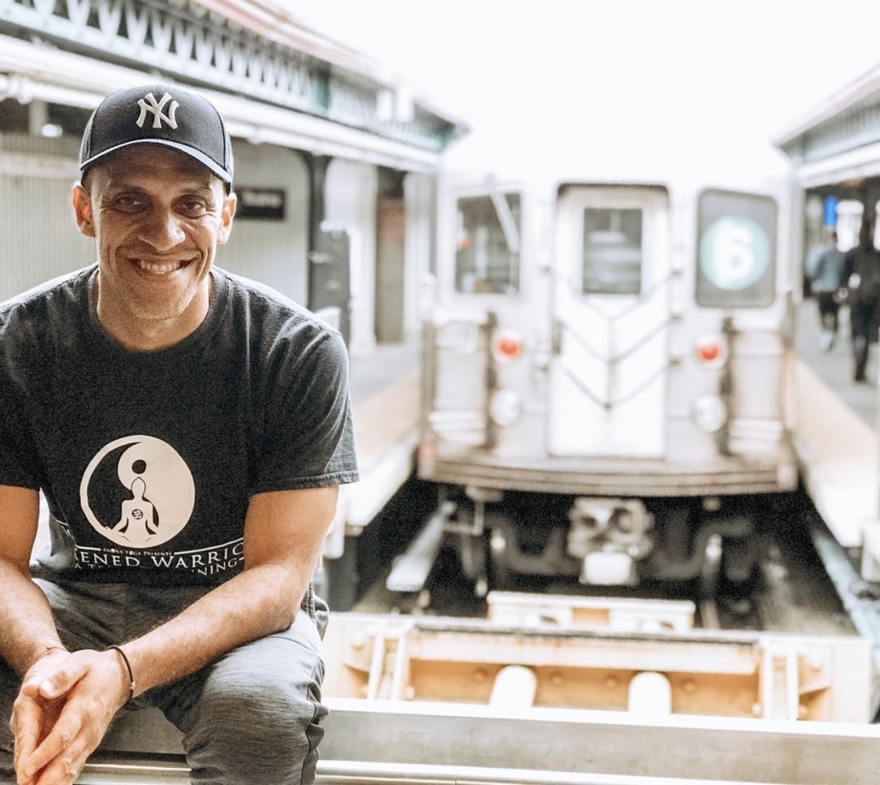 Smiling man wearing a New York Yankees cap and black T-shirt, sitting on a platform at a subway station with train behind him.
