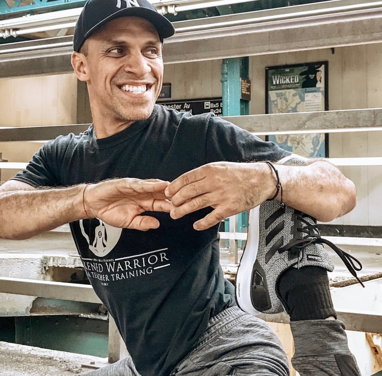 Smiling man stretching outdoors, wearing a black cap and T-shirt, sitting on a bench with a leg brace, in an urban setting.