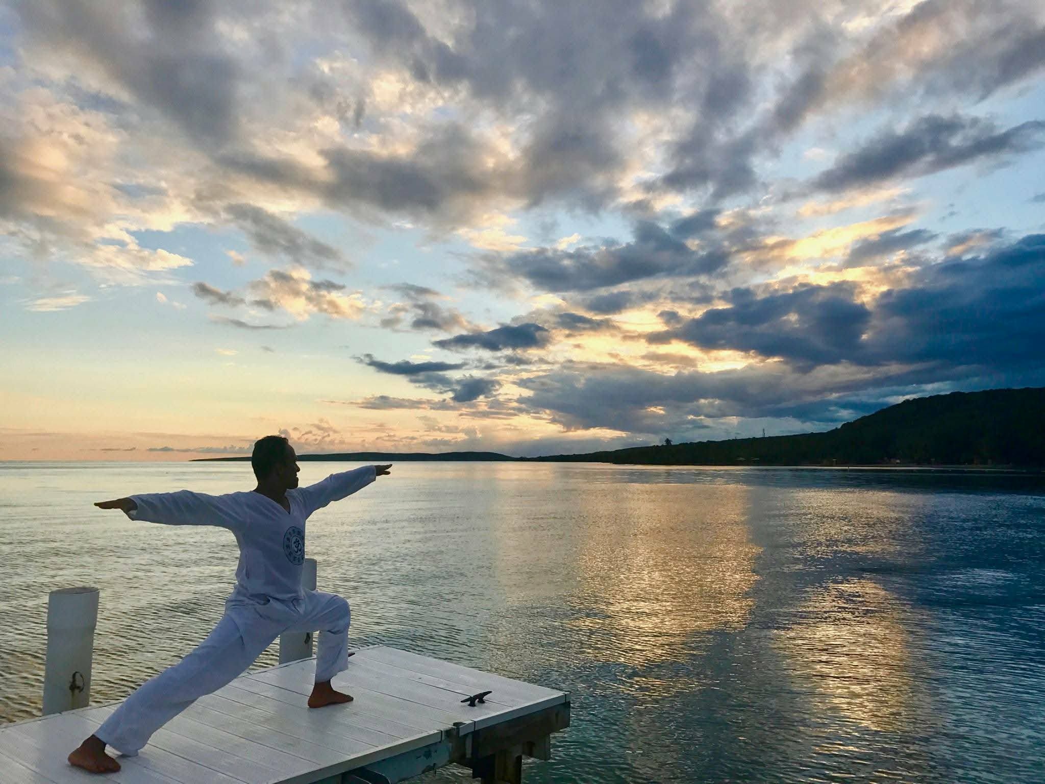 Person practicing yoga on a dock by water during sunset, with arms extended and clouds in the sky.