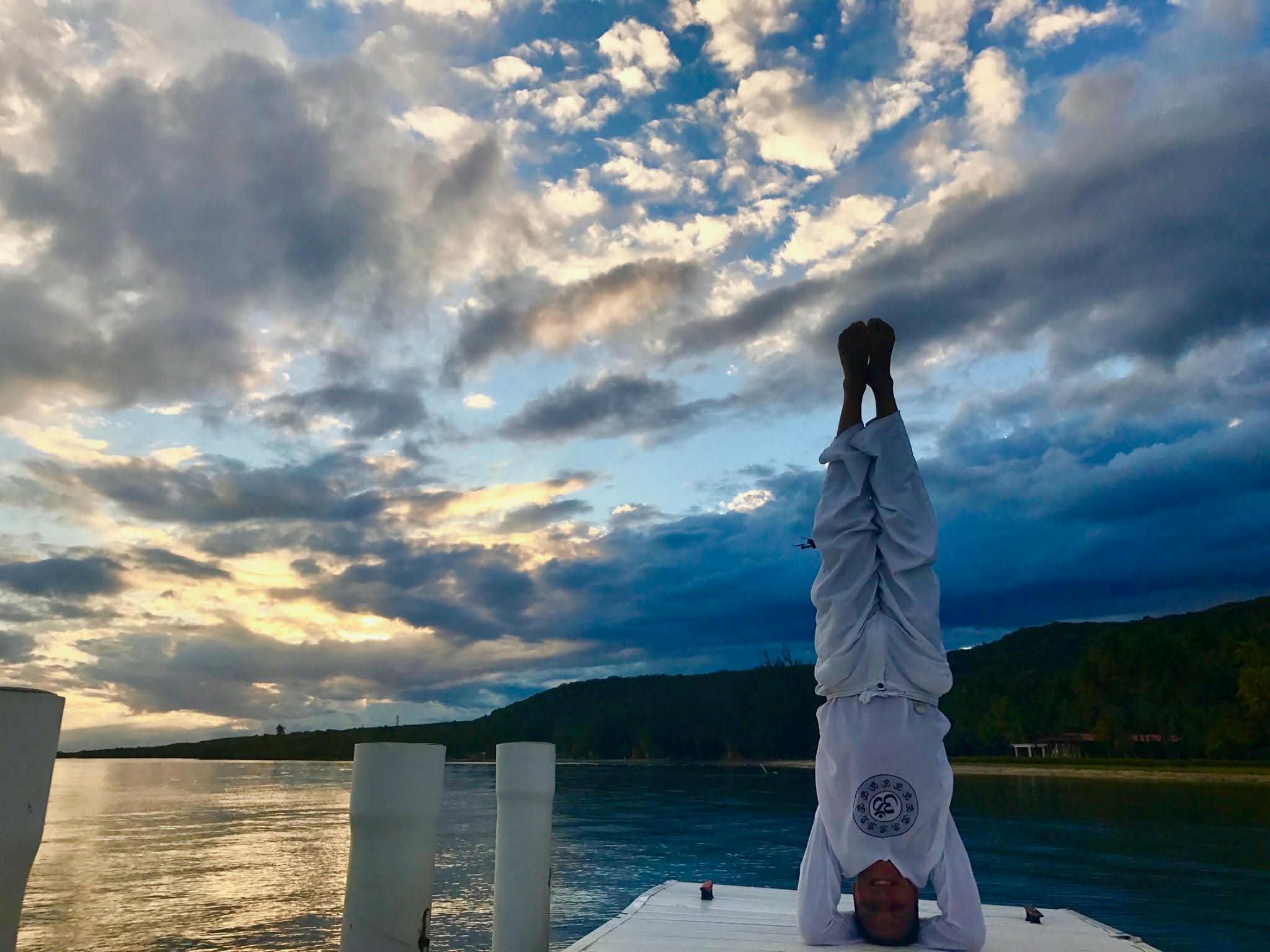 Person in white clothing standing on a boat with arms raised, sky with clouds and water in the background.