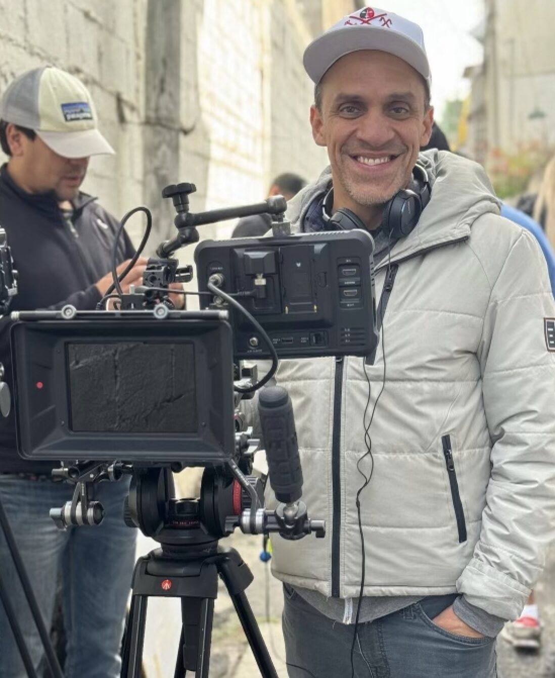 Man smiling in front of camera equipment on a city street, wearing a white jacket and cap.