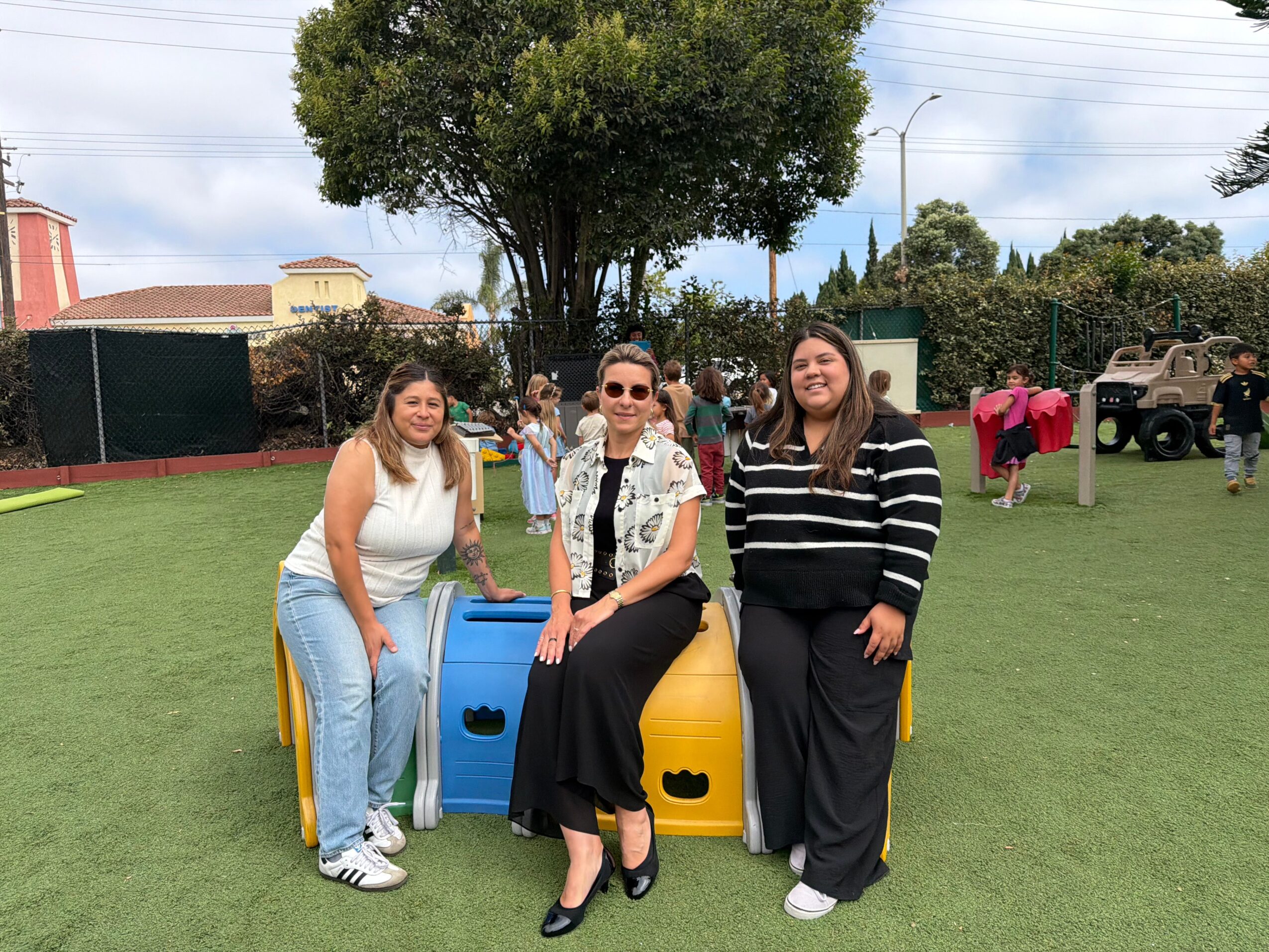 Three women sitting on a colorful playground structure outdoors, with a tree and other children in the background.