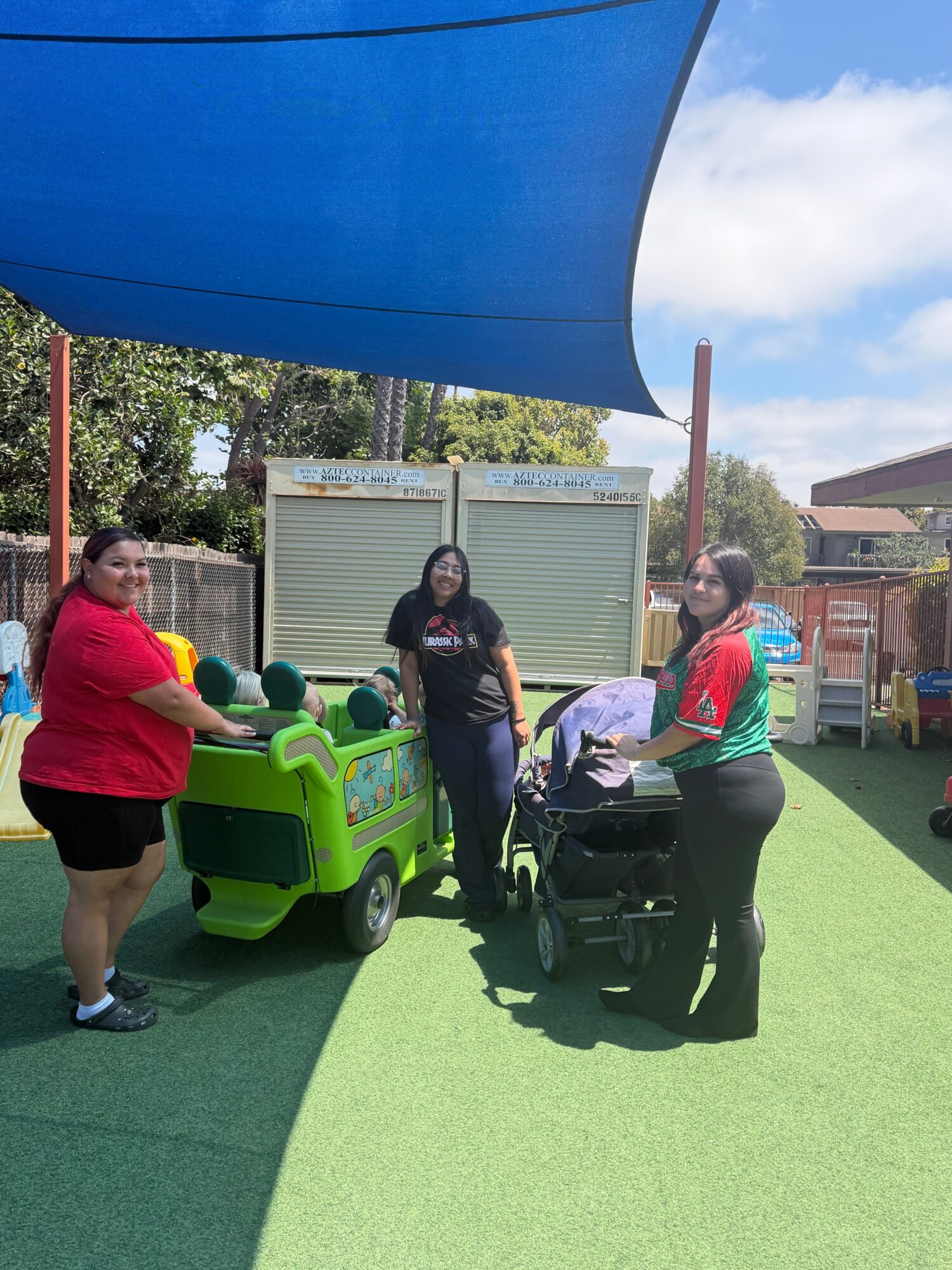 Person in red shirt and black shorts interacts with a small green vehicle, with another person in a green jacket nearby, outdoors.