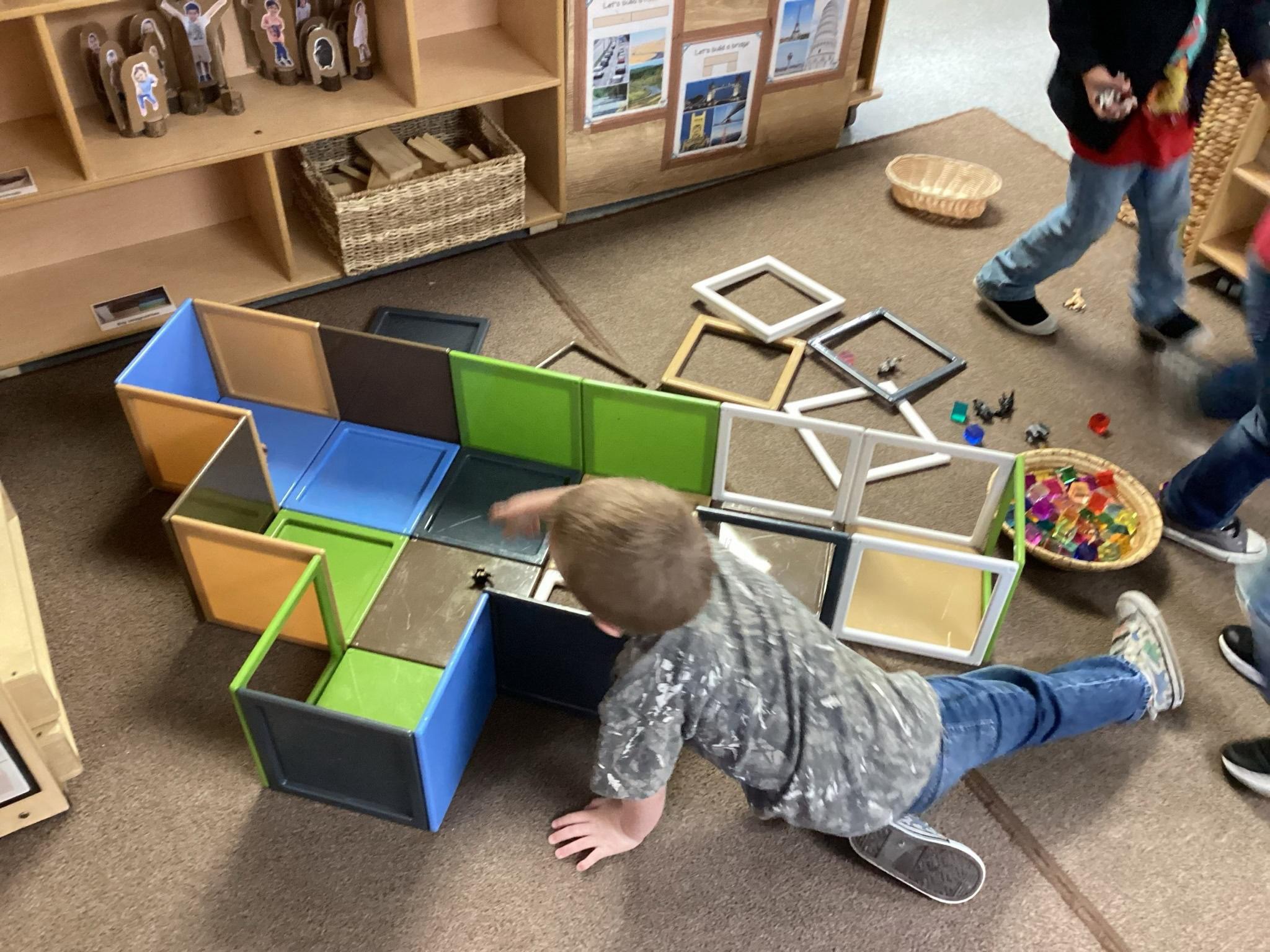 Child lying on the floor playing with colorful modular blocks in a classroom, with other children and shelves in the background.