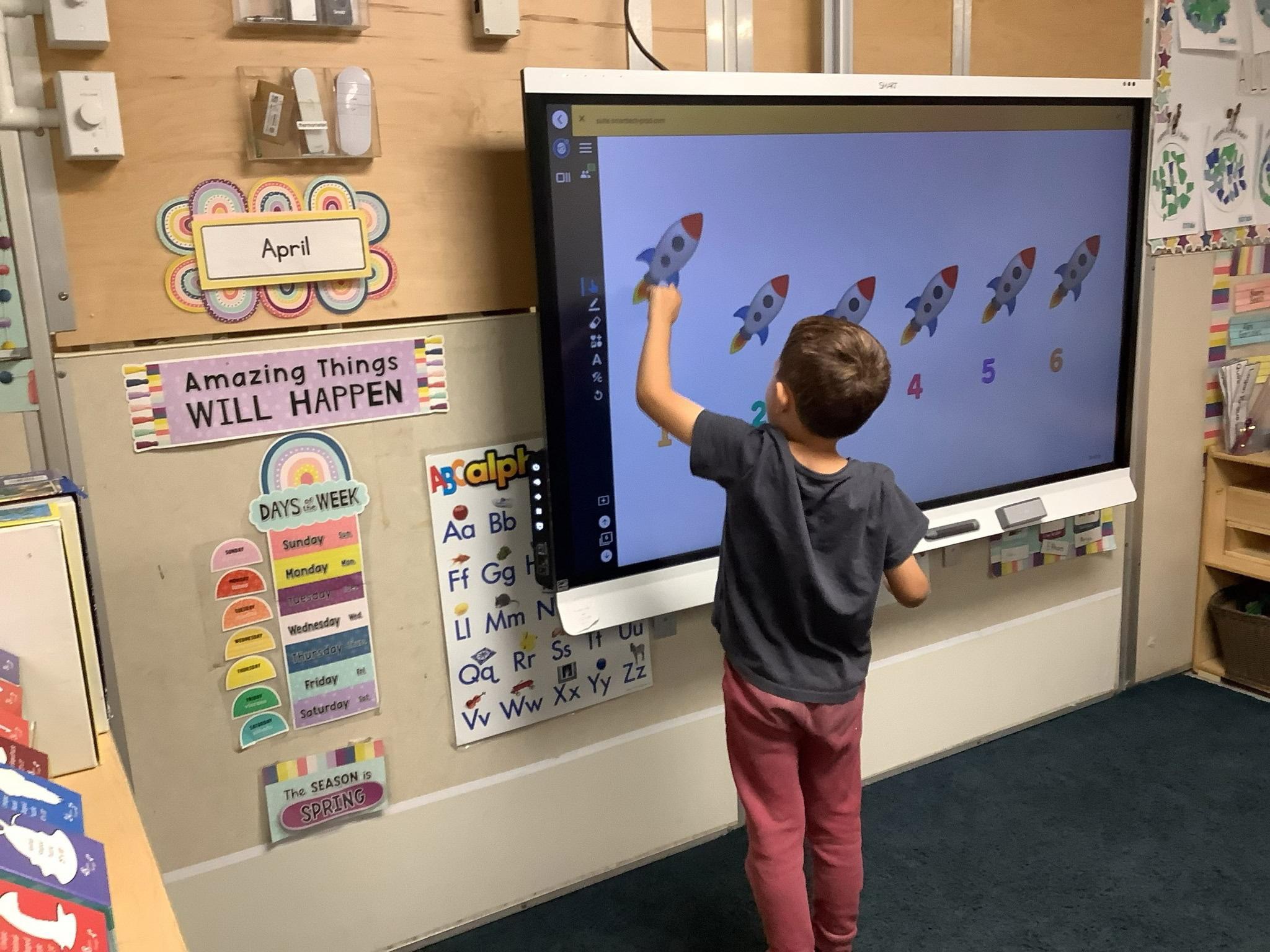 Child interacts with a digital screen displaying rockets, in a classroom with educational posters and a calendar, standing on a dark carpet.