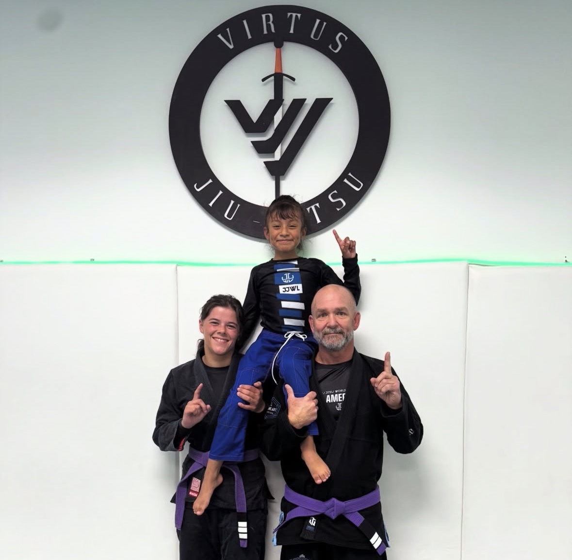 Three people in martial arts uniforms pose in front of a Virtus Jiu Jitsu sign, with one person sitting on another's shoulders, all smiling and showing a number one gesture.