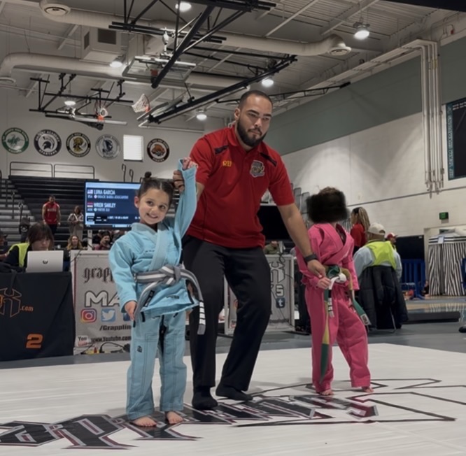 Martial arts instructor with two children in a gym, one child in a blue gi, the other in pink, practicing martial arts.