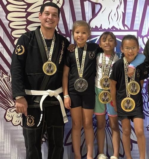 Four children and one adult in martial arts uniforms with medals, standing in front of a colorful background.