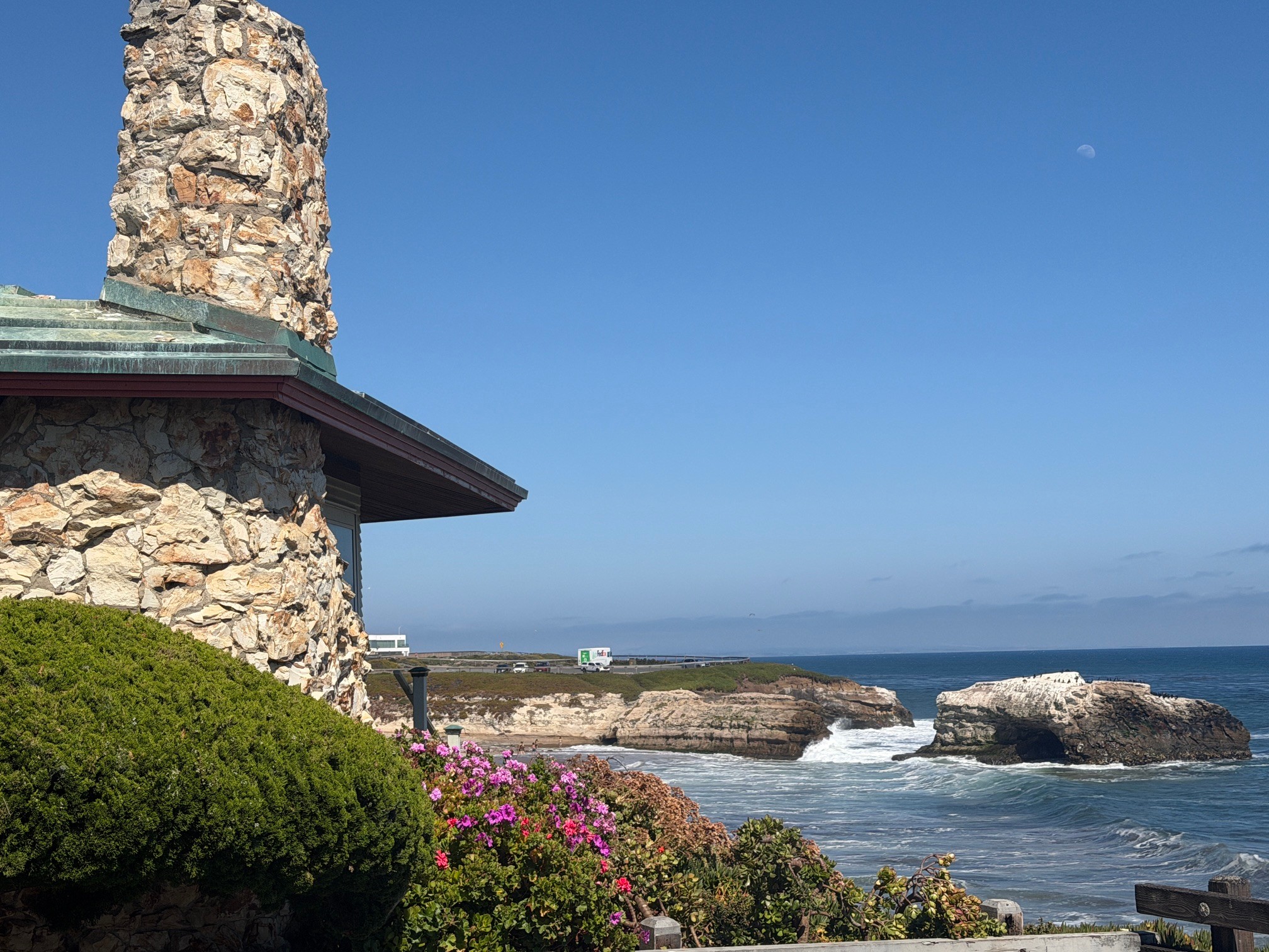 Stone building with chimney near ocean, green bushes, pink flowers, and rocky shoreline under blue sky.