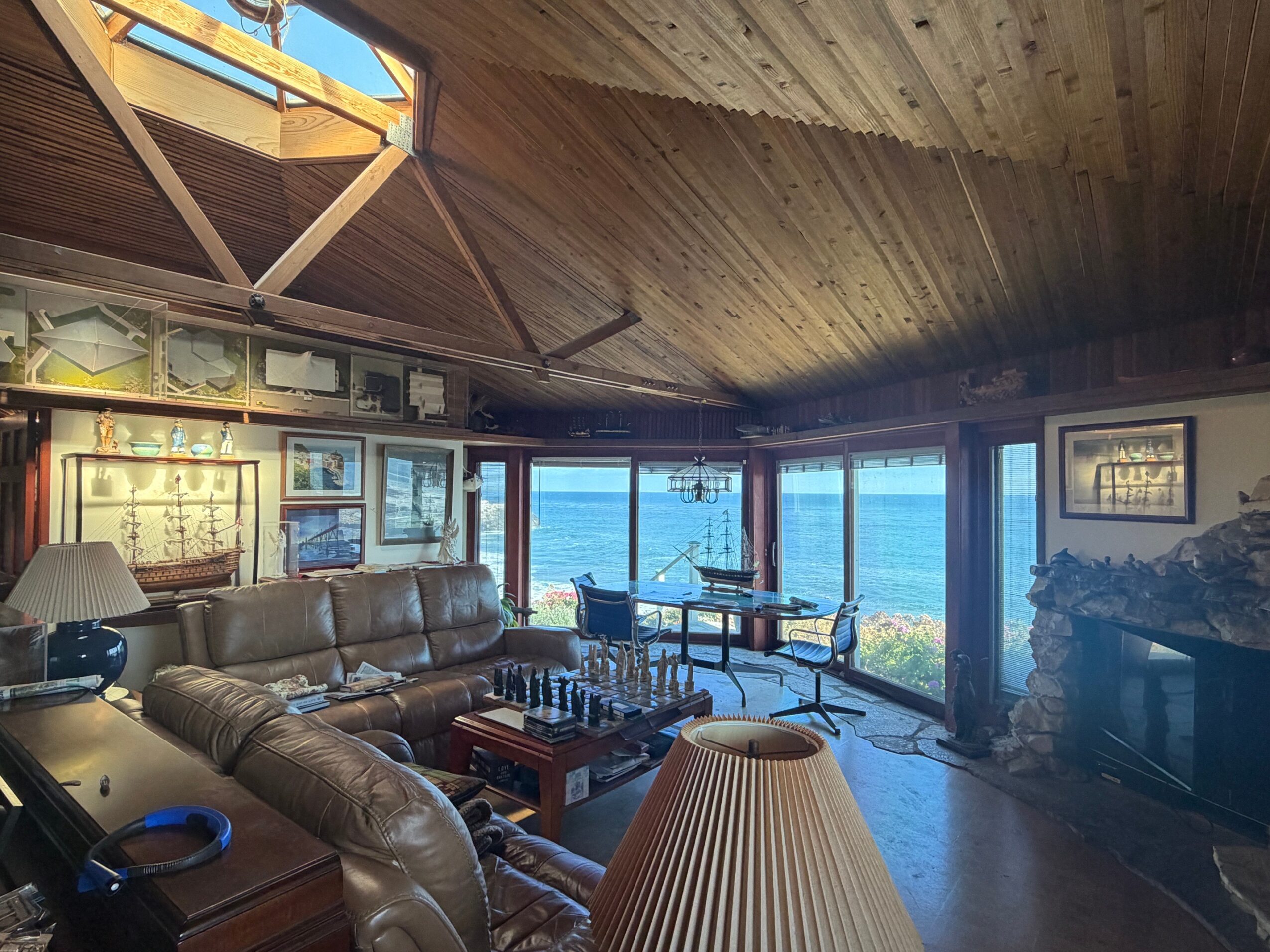 Living room with large windows overlooking the ocean, wooden ceiling, leather sofas, and framed pictures on the wall.