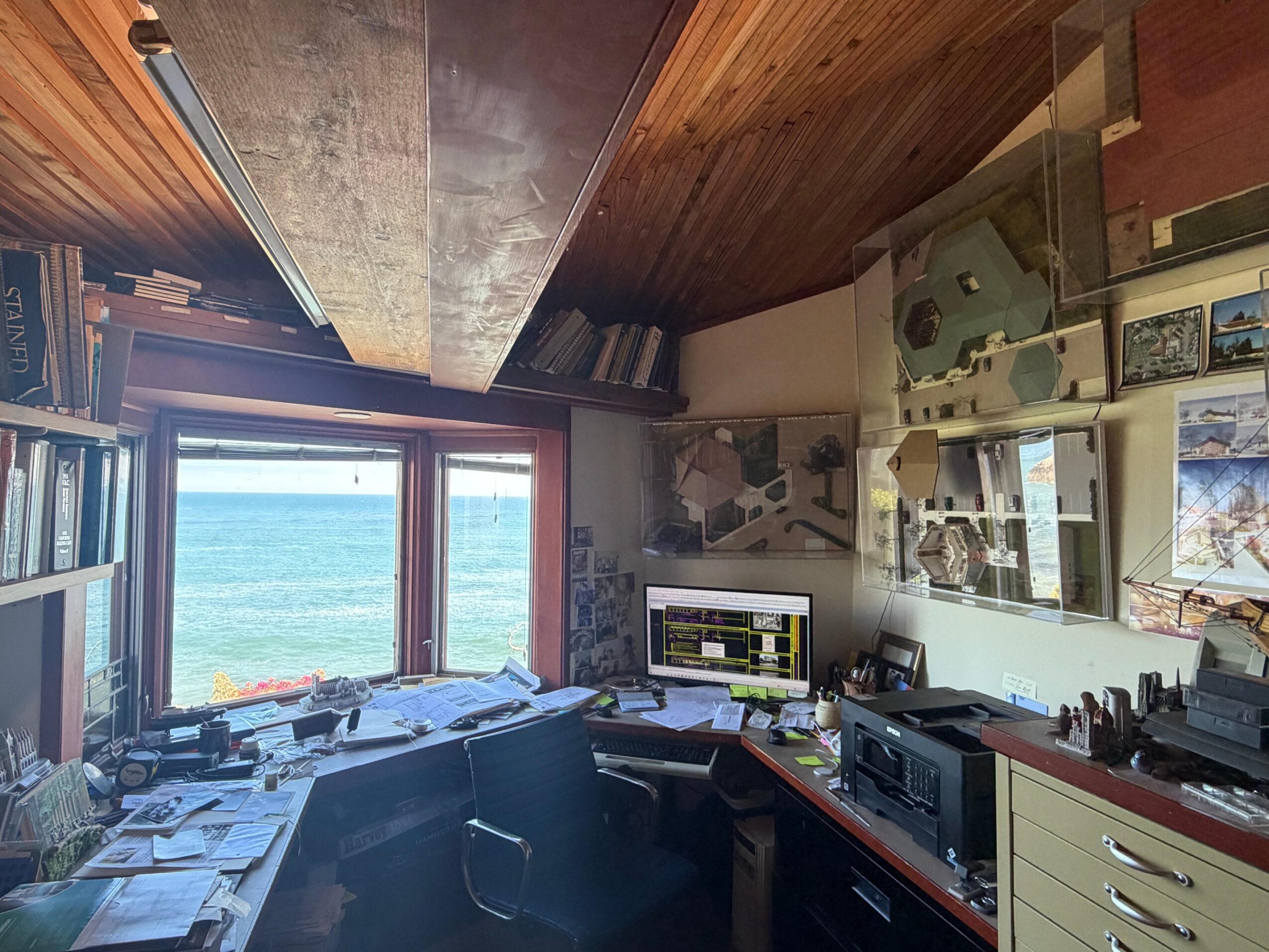 Room with a large window showing a body of water, cluttered desk, bookshelves, and various artwork on the walls.
