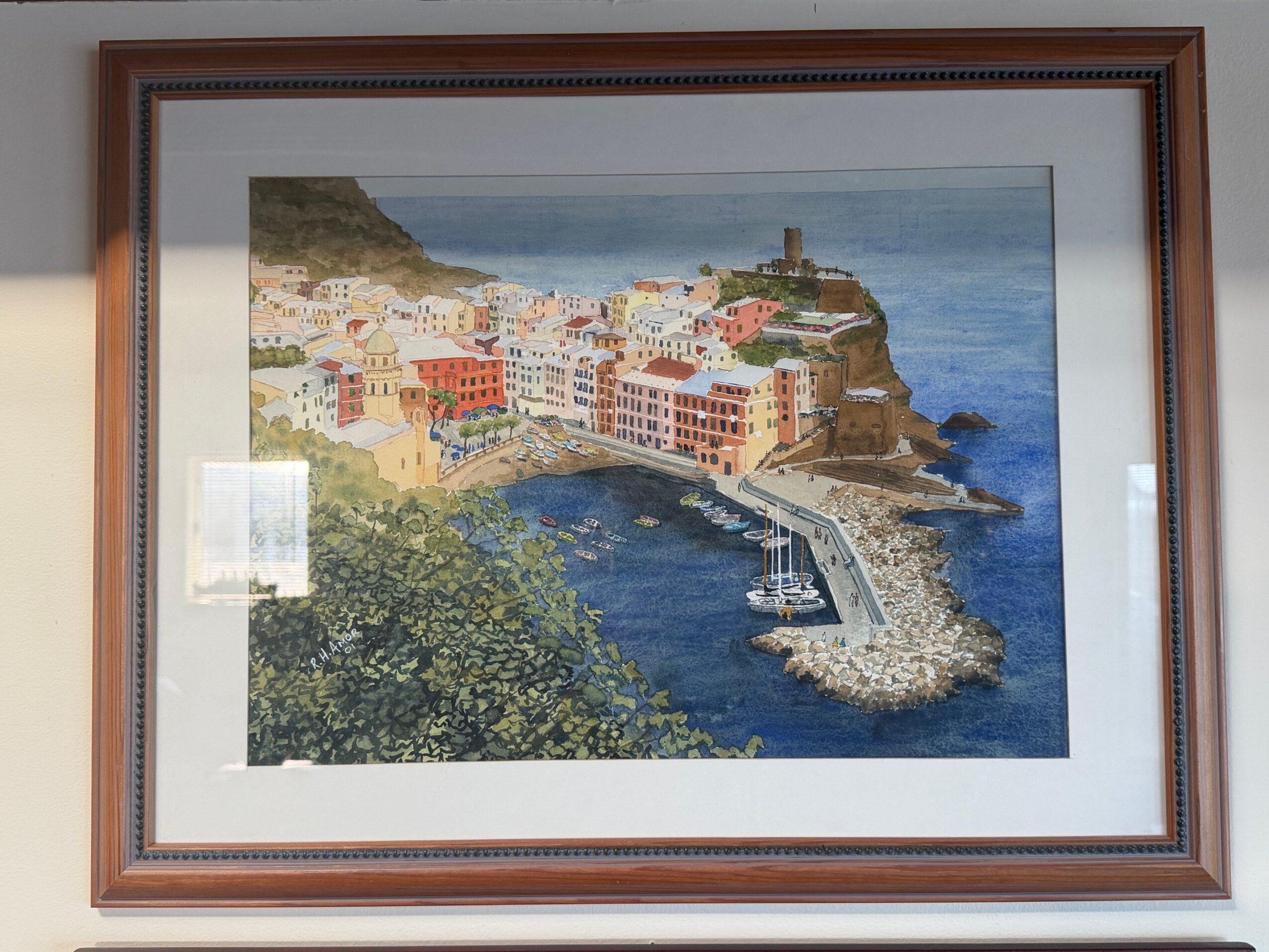 Framed painting of a coastal town with colorful buildings, a harbor, and a lighthouse on a rocky hill.