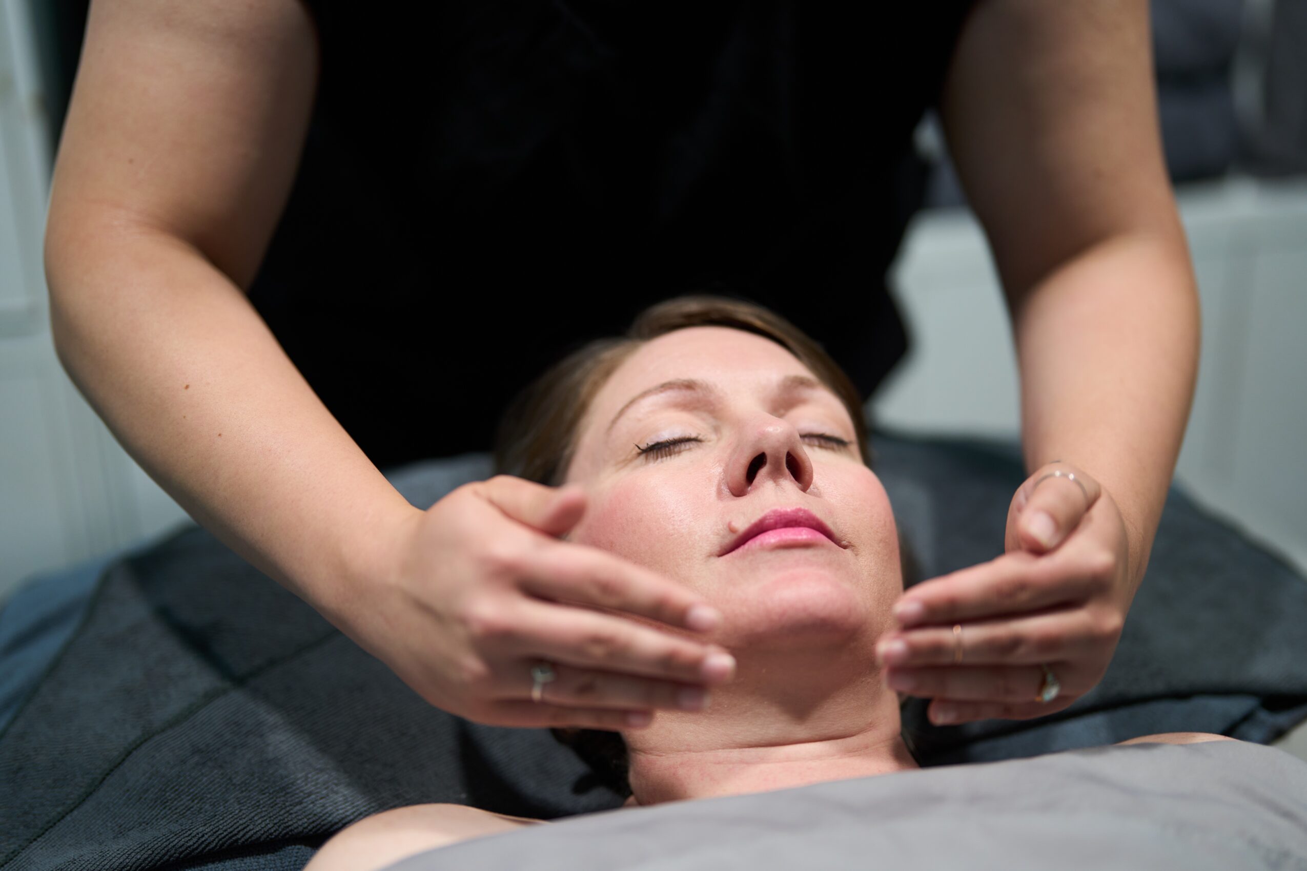 Person receiving a facial massage with hands on face, eyes closed, lying down, in a spa or treatment setting.