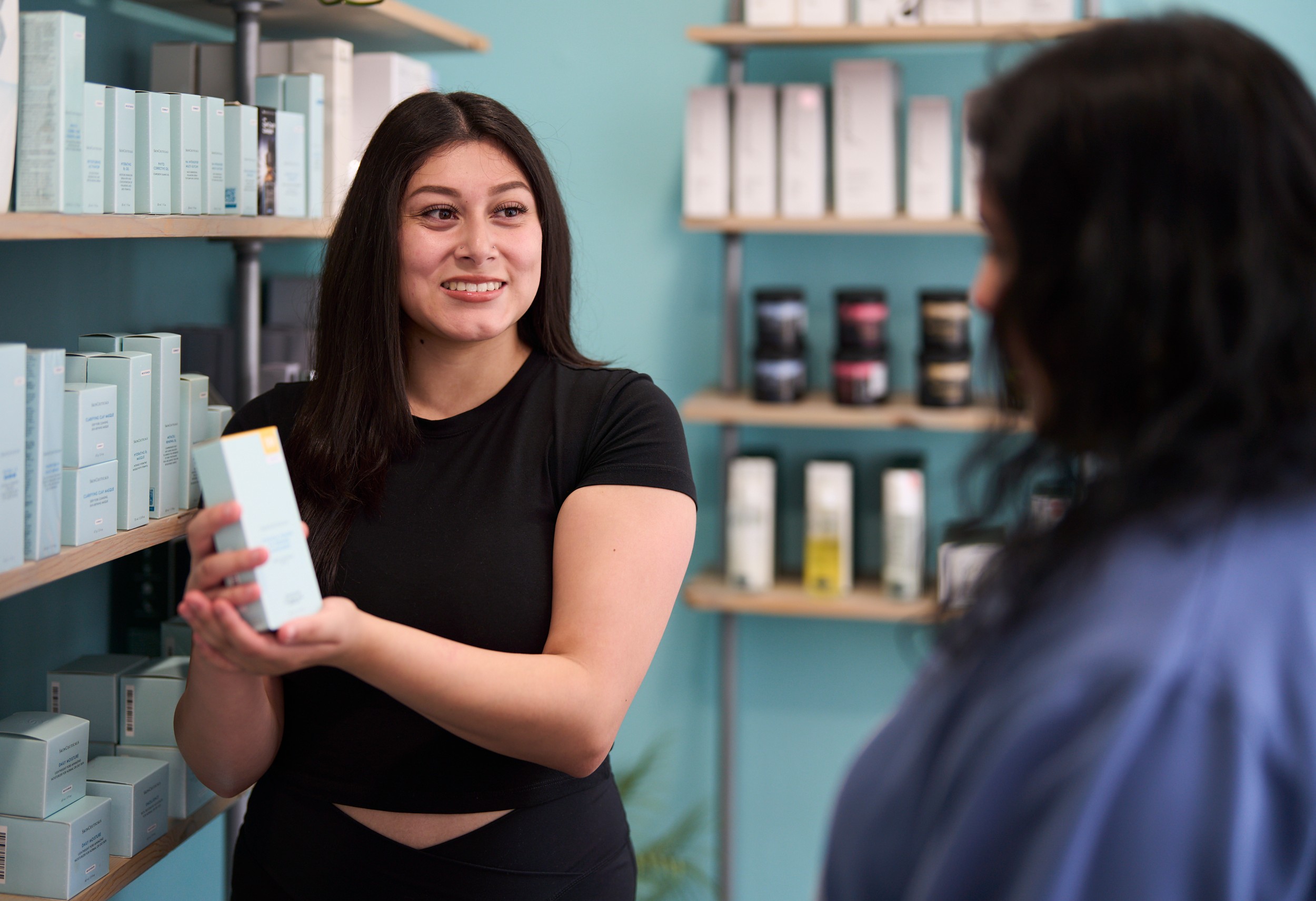 Two women in a store aisle, one smiling and holding a product, the other facing away, shelves with products behind them.