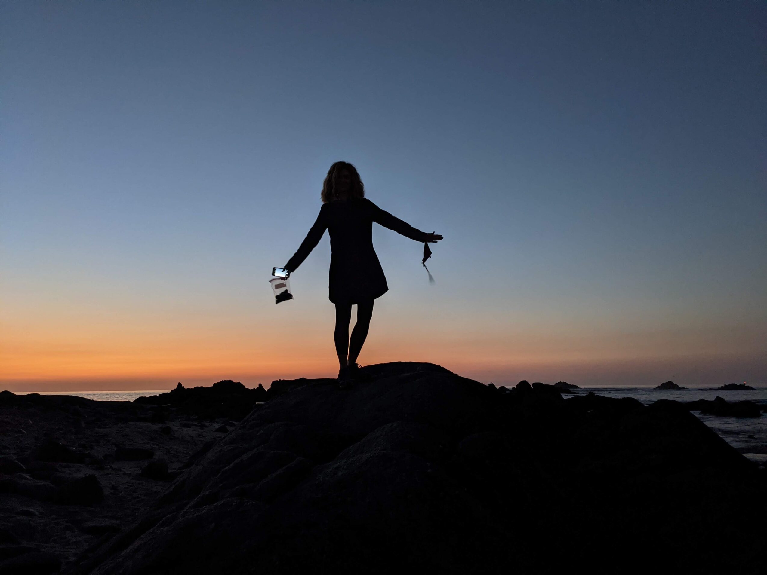 Silhouette of a person standing on a rock at sunset, holding a lantern and a scarf, with a colorful sky.