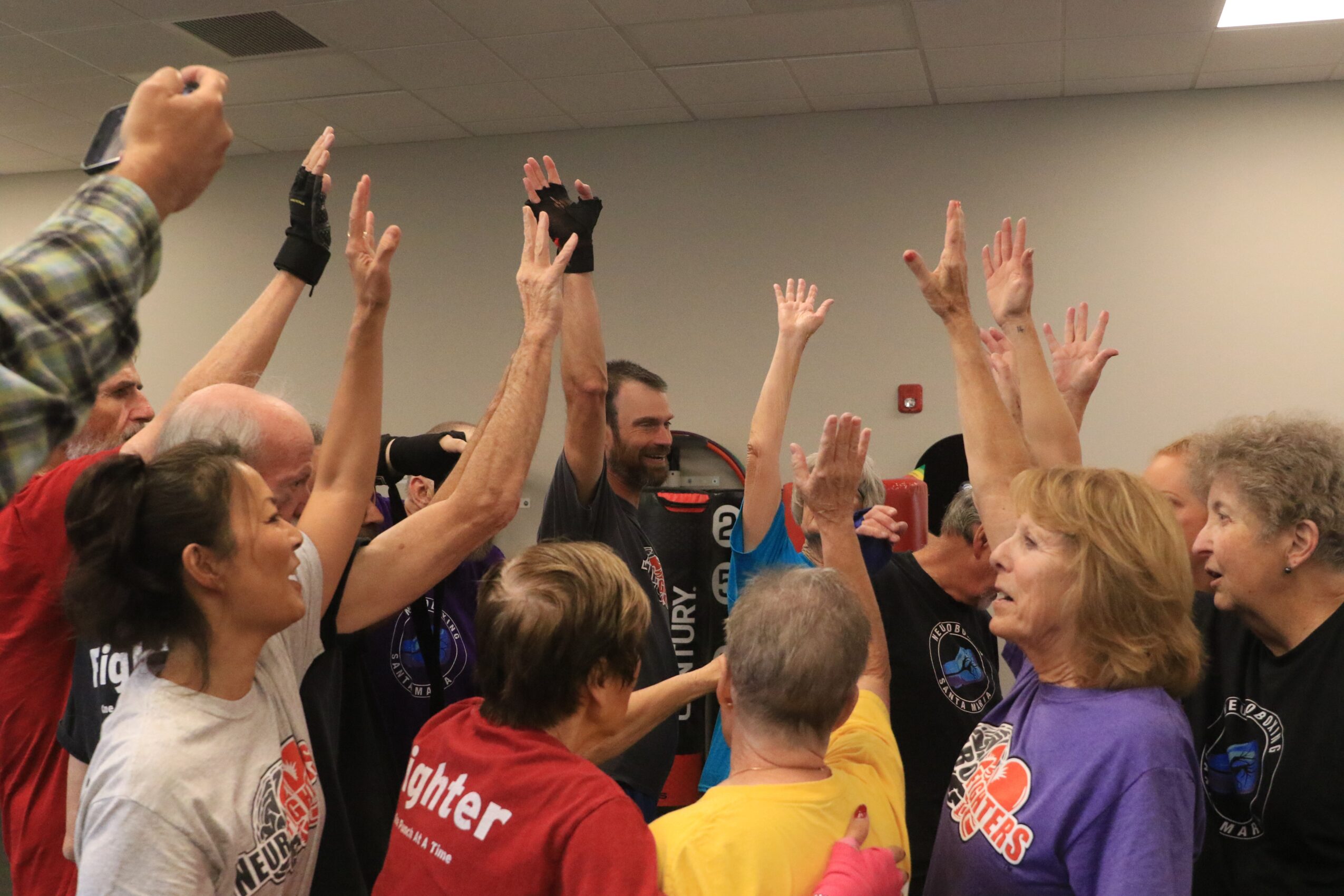 Group of people raising hands in a room with a plain wall and ceiling tiles.