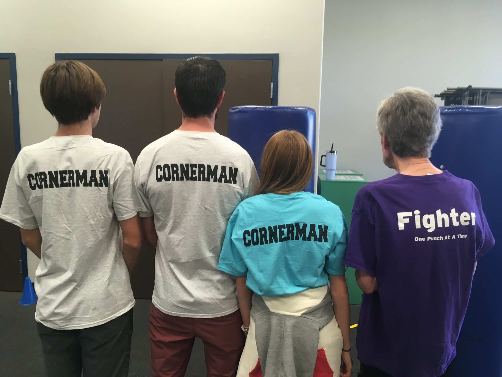 Four people standing with their backs to the camera, two wearing white shirts, one in a blue shirt, and one in a purple shirt, in front of a screen.