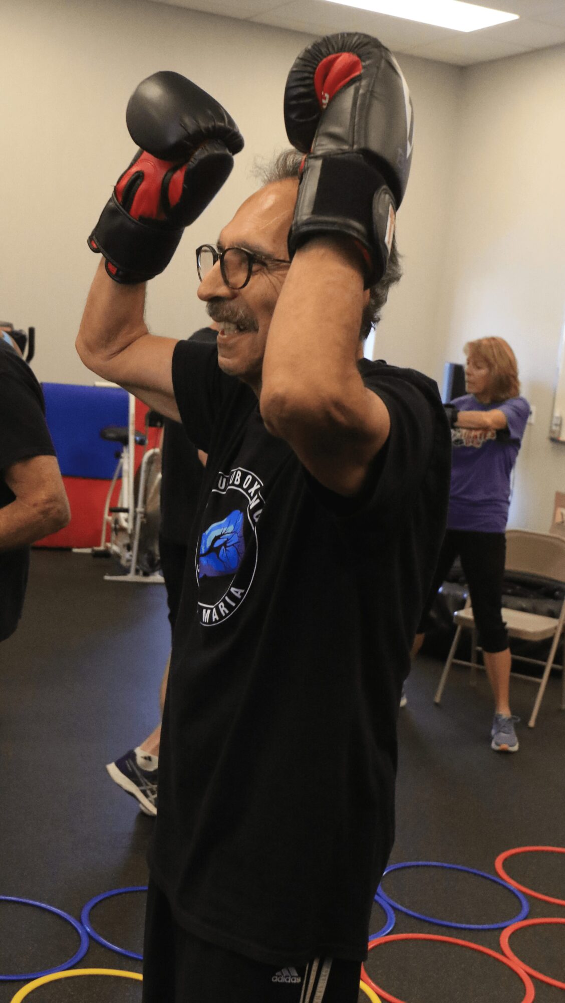 Man wearing boxing gloves and glasses, raising arms in a gym with people in the background.
