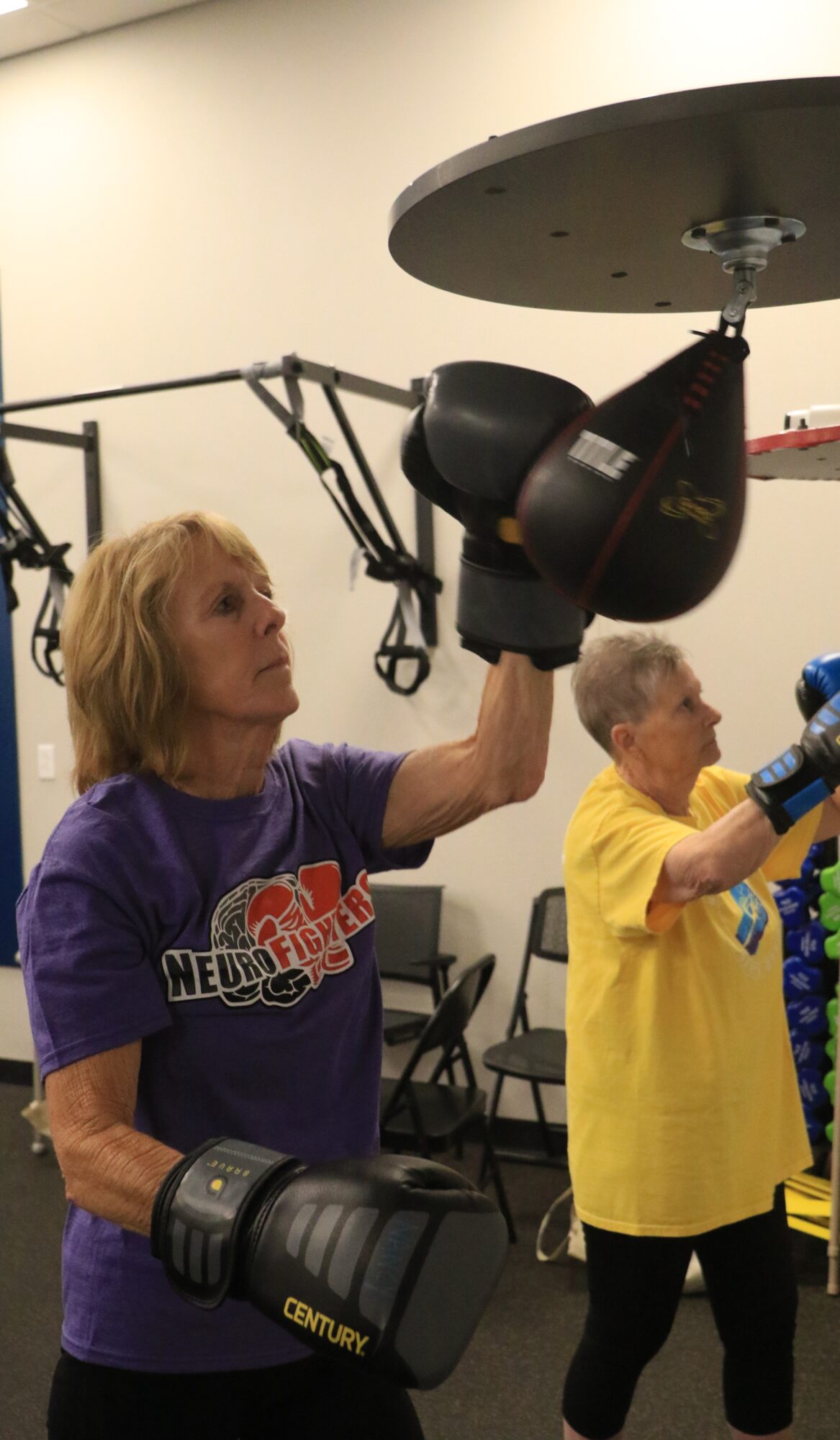 Two women in a gym using punching bags, one in a purple shirt and the other in a yellow shirt, with gym equipment in background.