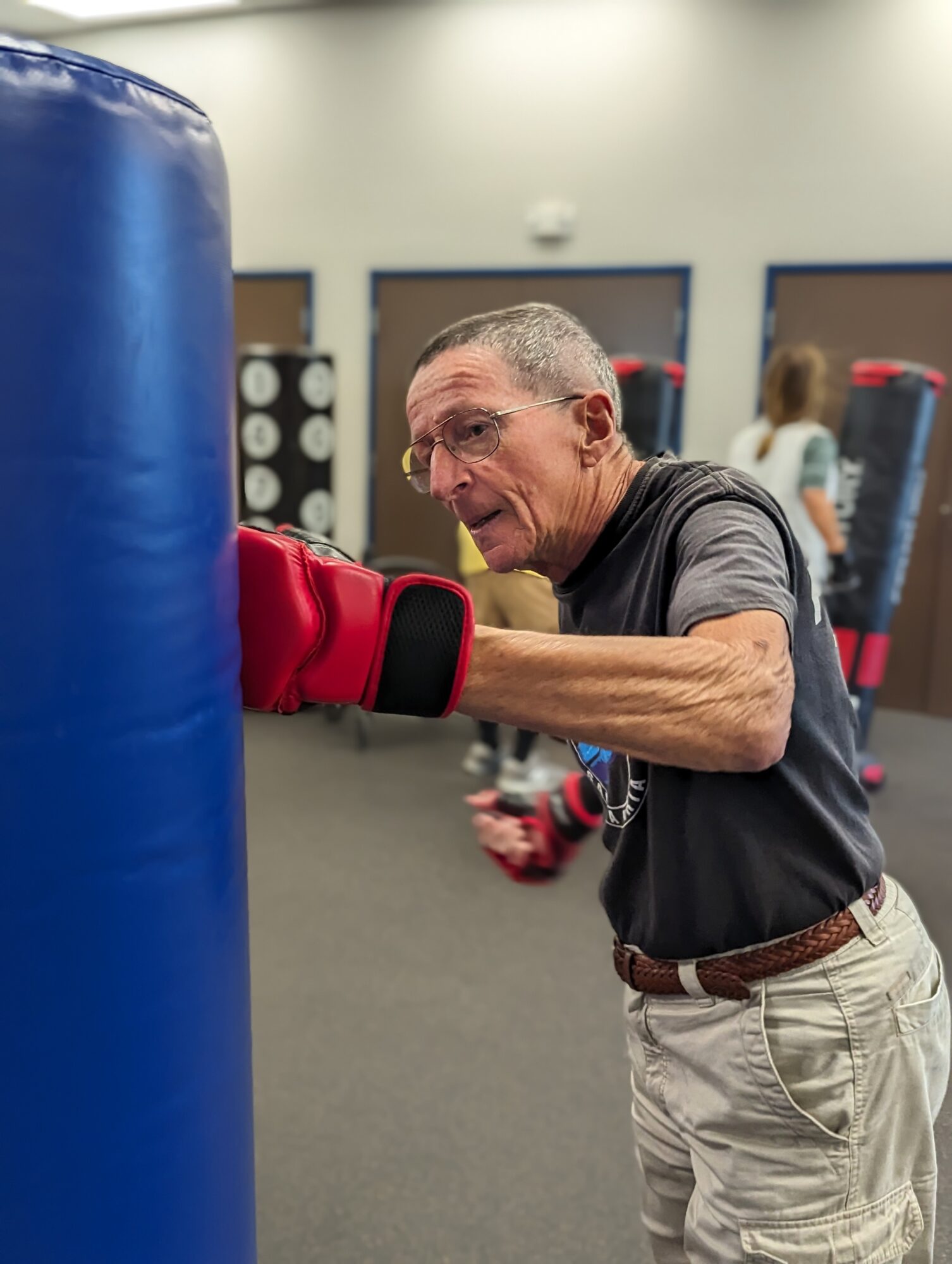 Elderly man wearing glasses and a black shirt punches a blue punching bag in a gym.