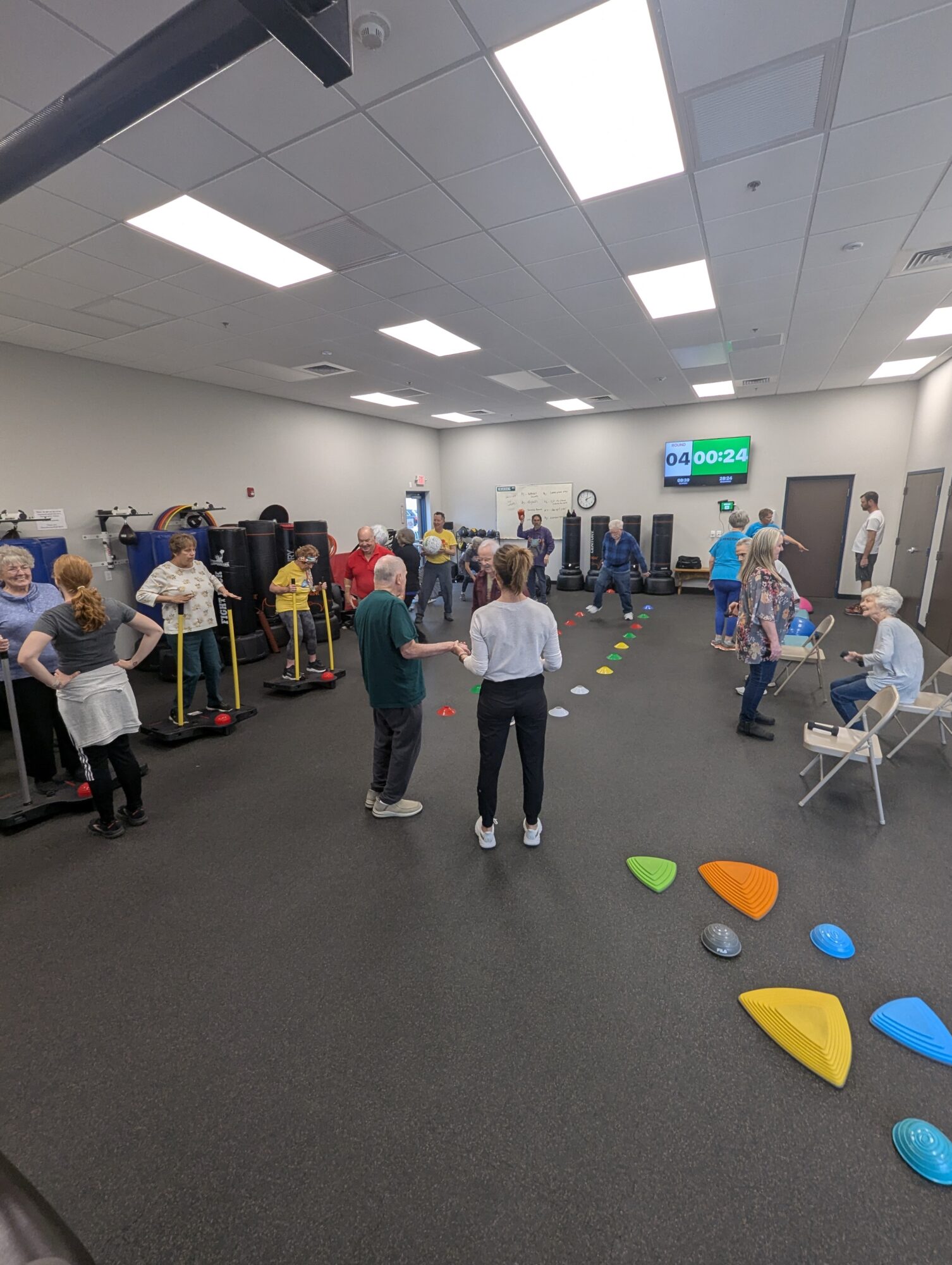 People in a room with chairs, colorful stepping stones, and a digital clock on the wall, engaging in activities.