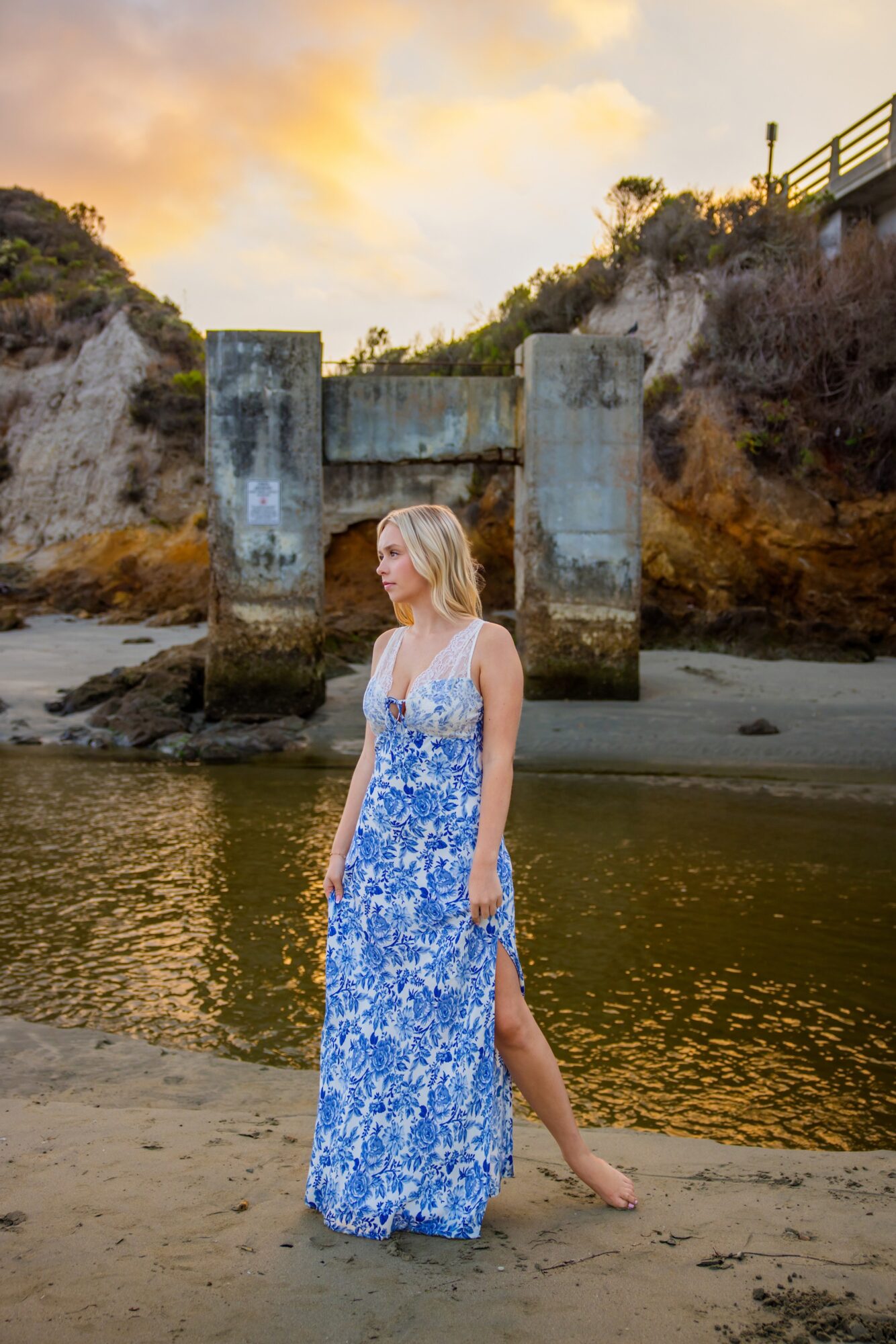 Woman in a blue and white floral dress standing on a beach near water with a bridge and trees in the background.