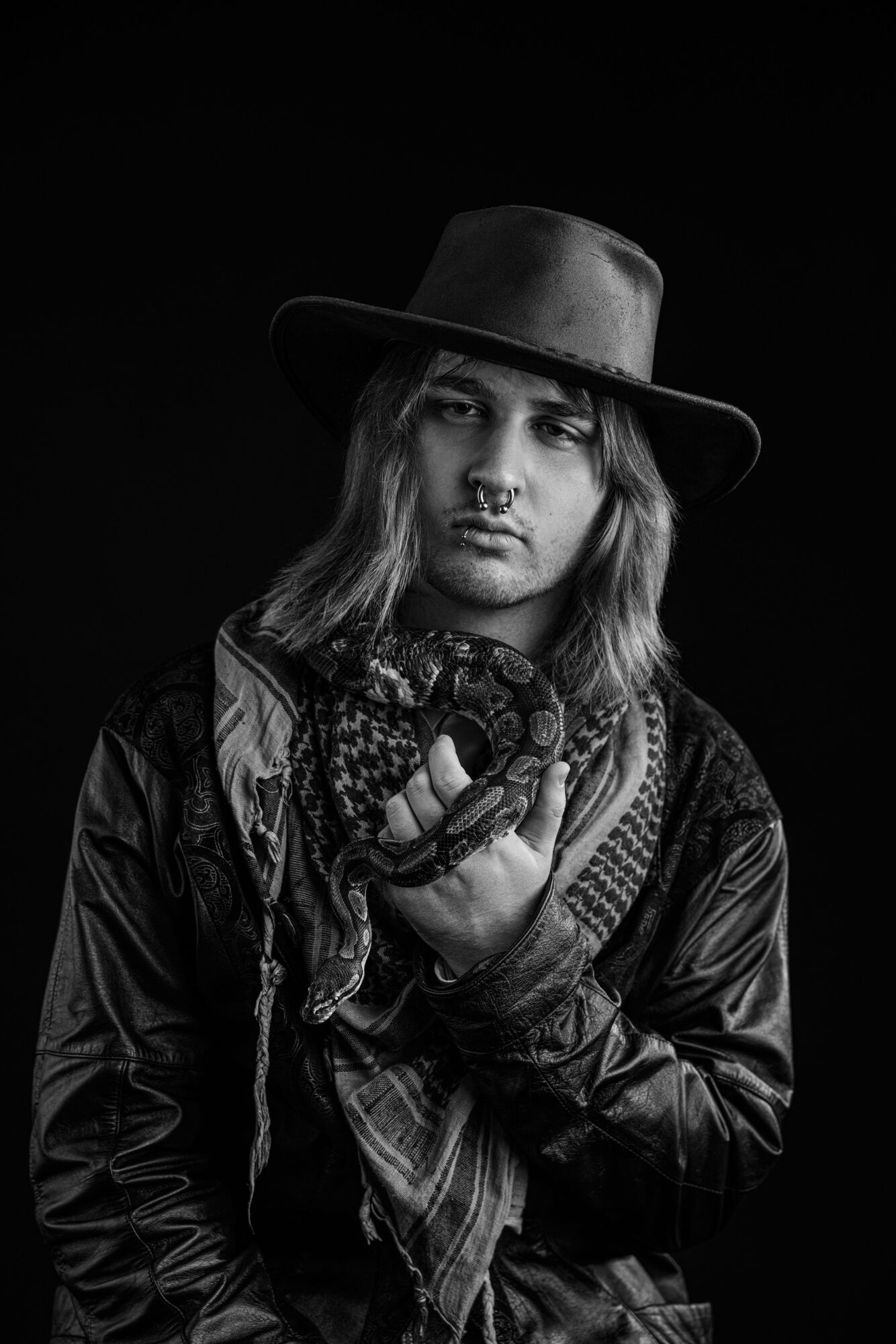 Person with long hair wearing a wide-brimmed hat, scarf, and leather jacket, holding a camera, in black and white.
