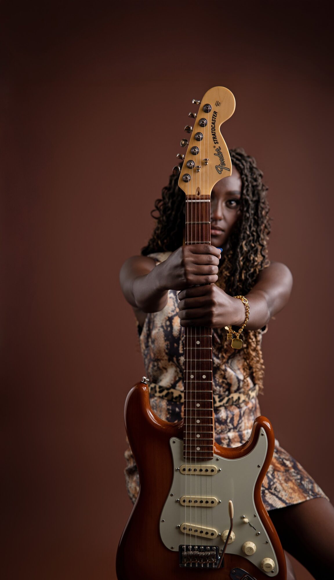 Person holding an electric guitar, partially obscured, with curly hair and a patterned outfit, against a dark background.