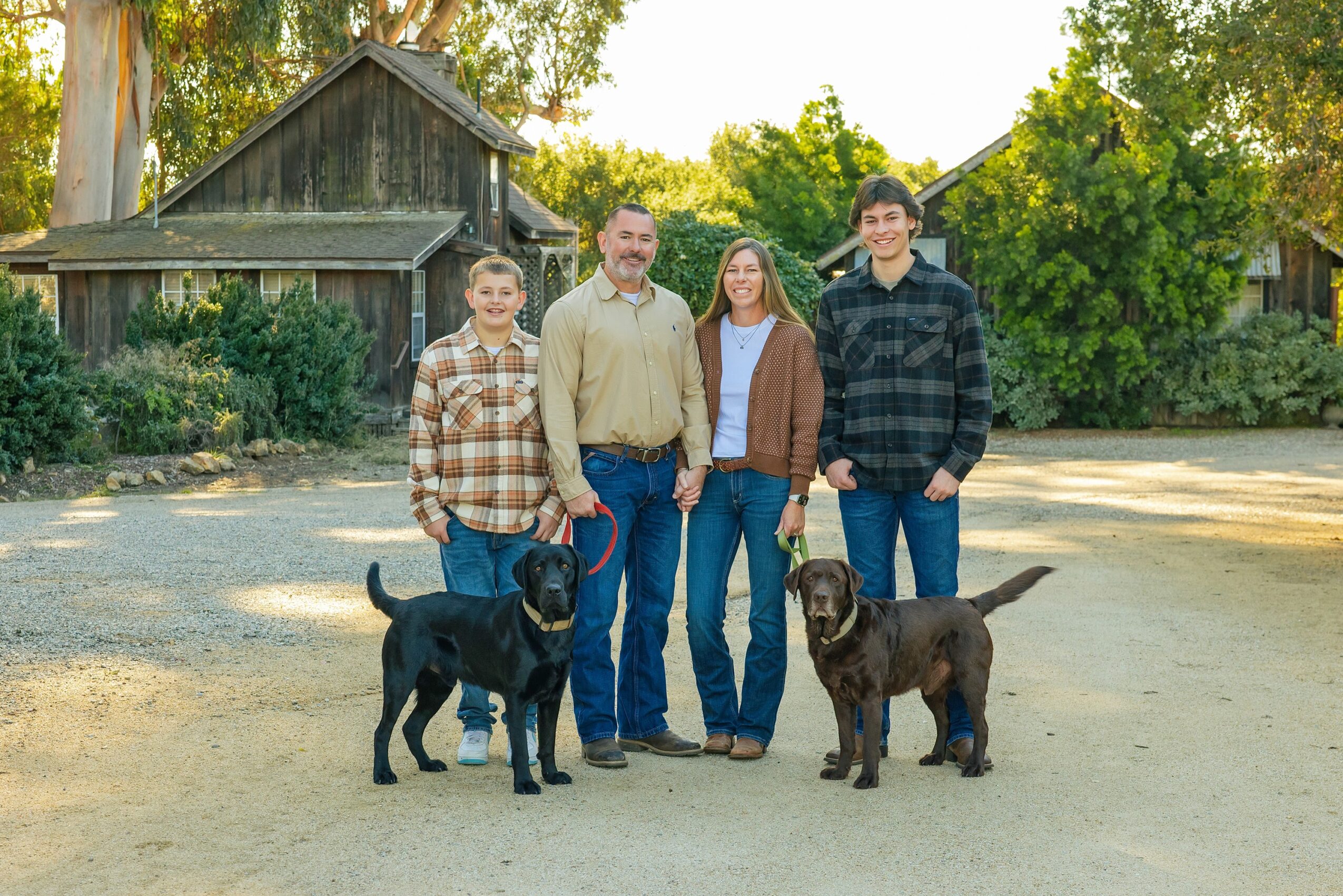 Four people stand outdoors with two dogs, trees and a wooden building in the background.