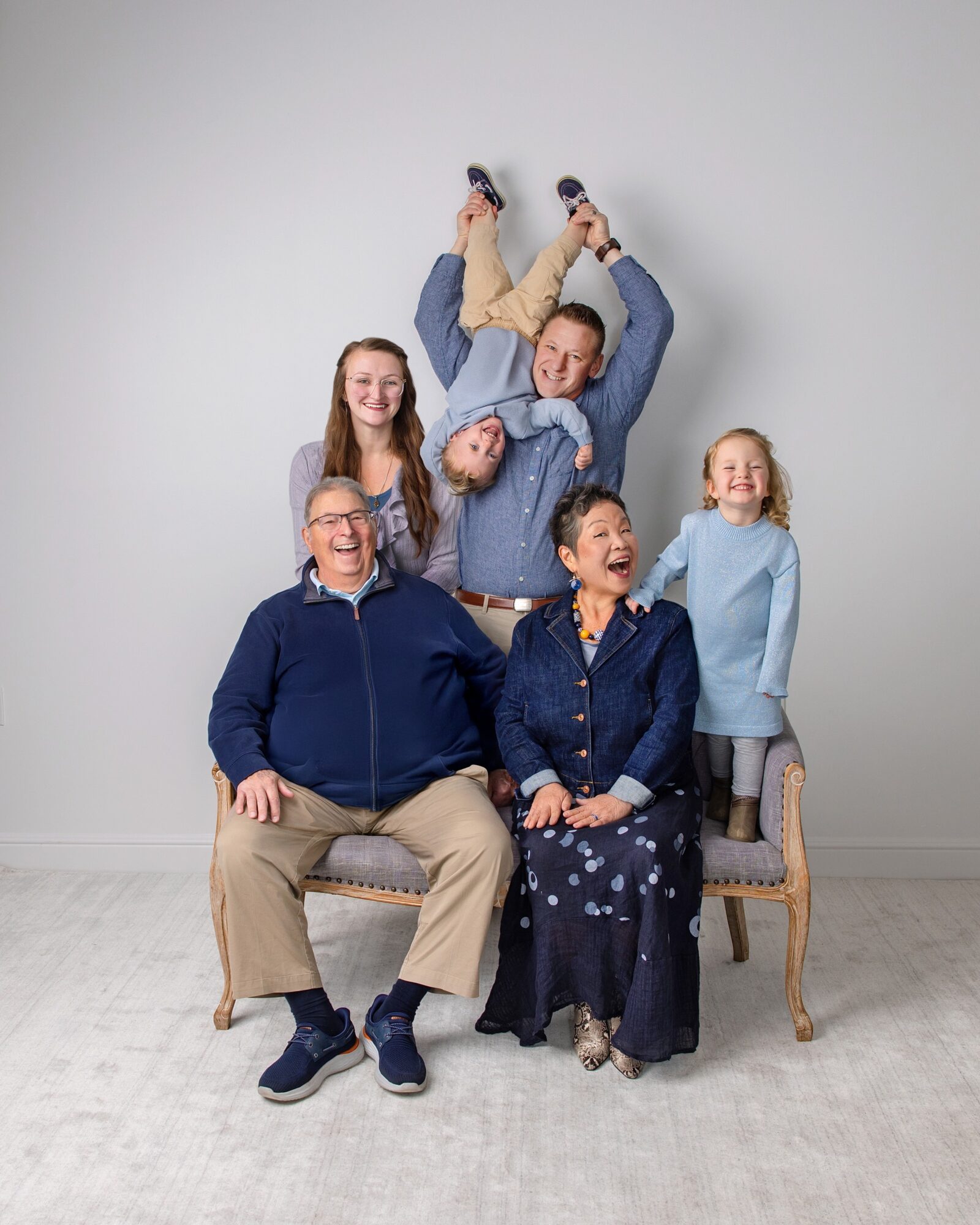 Group of six people, five women and one man, posing with one person upside down, smiling and laughing, against a plain wall.