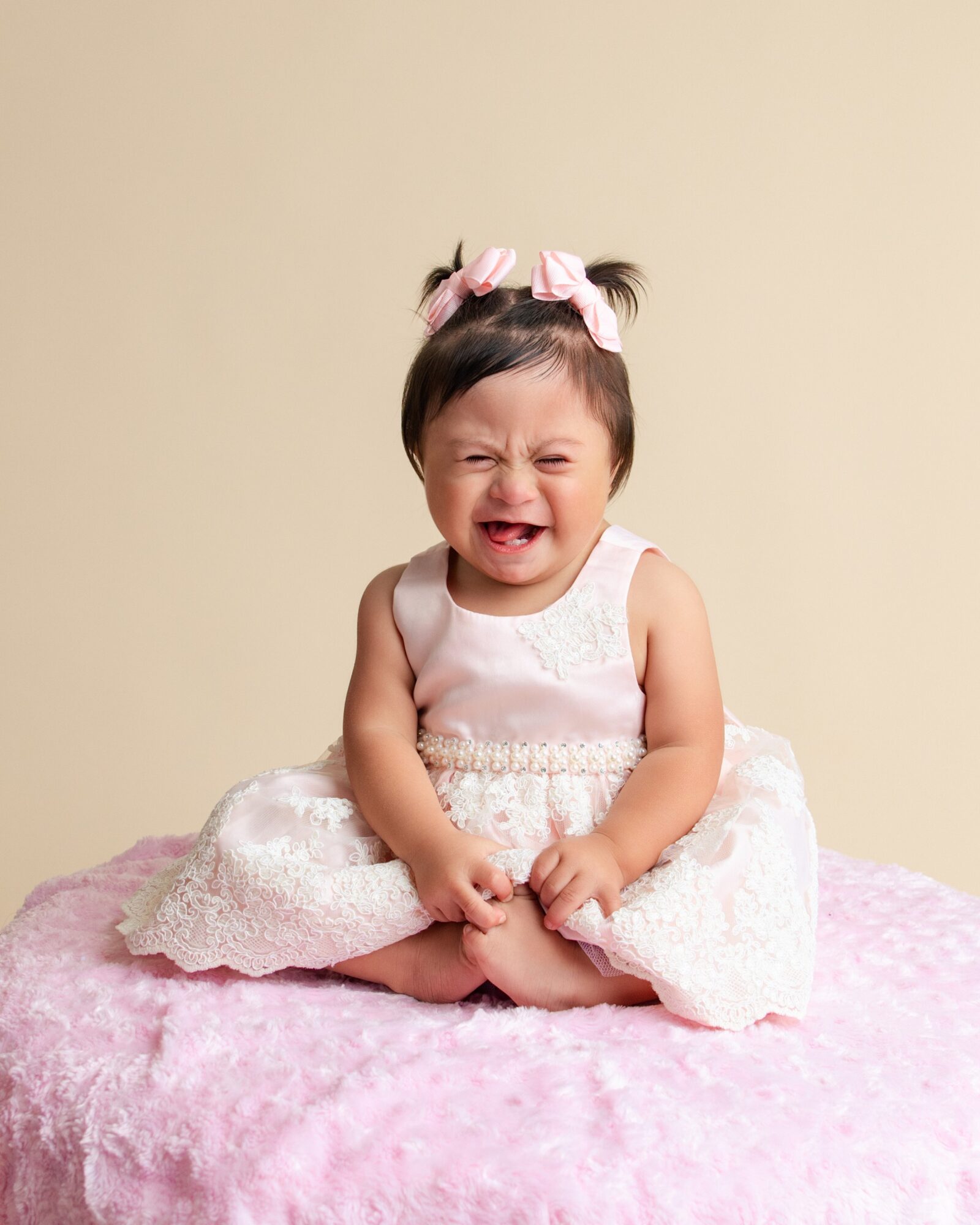 Smiling baby girl sitting on pink blanket, wearing a white dress and pink bows in hair, against beige background.
