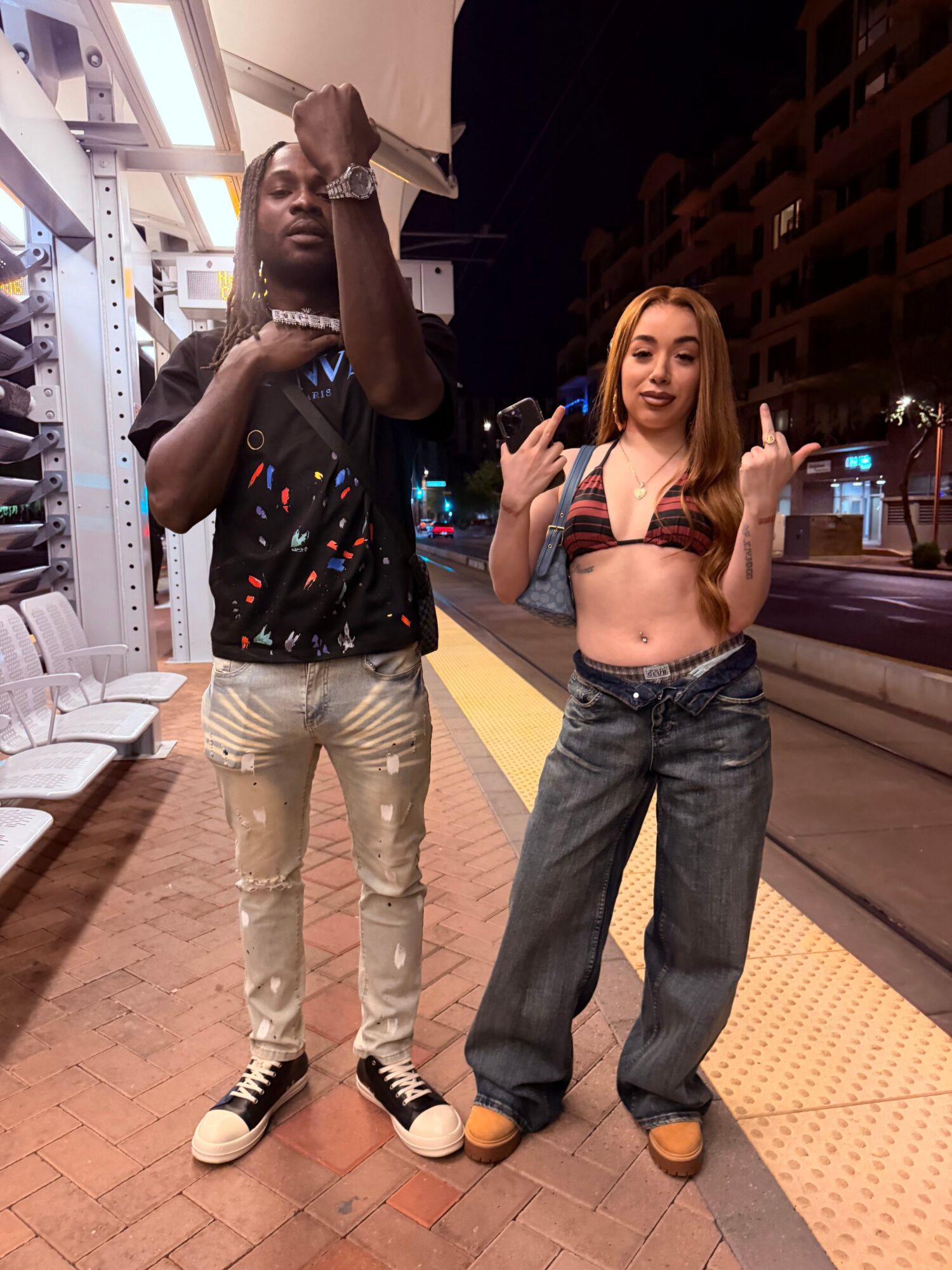 Two young women stand on a city sidewalk at night, one with long dark hair and the other with long blonde hair, both making hand gestures.
