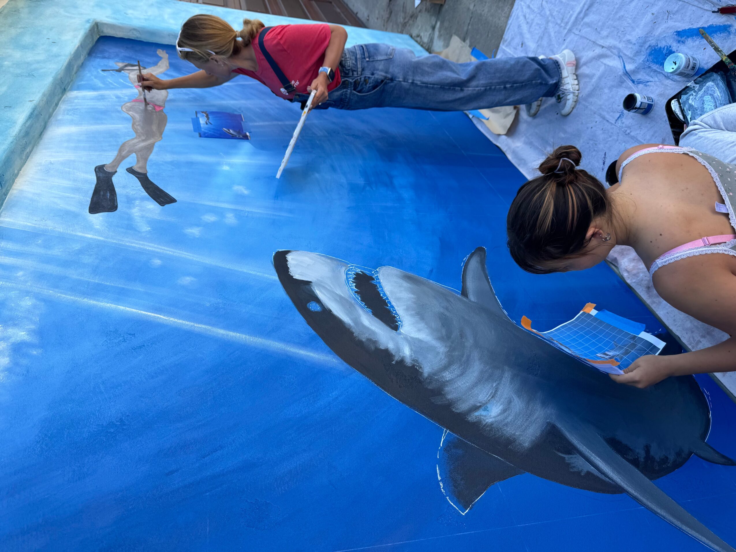 Two women interact with a painted shark on a pool wall, one holding a large shark model, the other painting.
