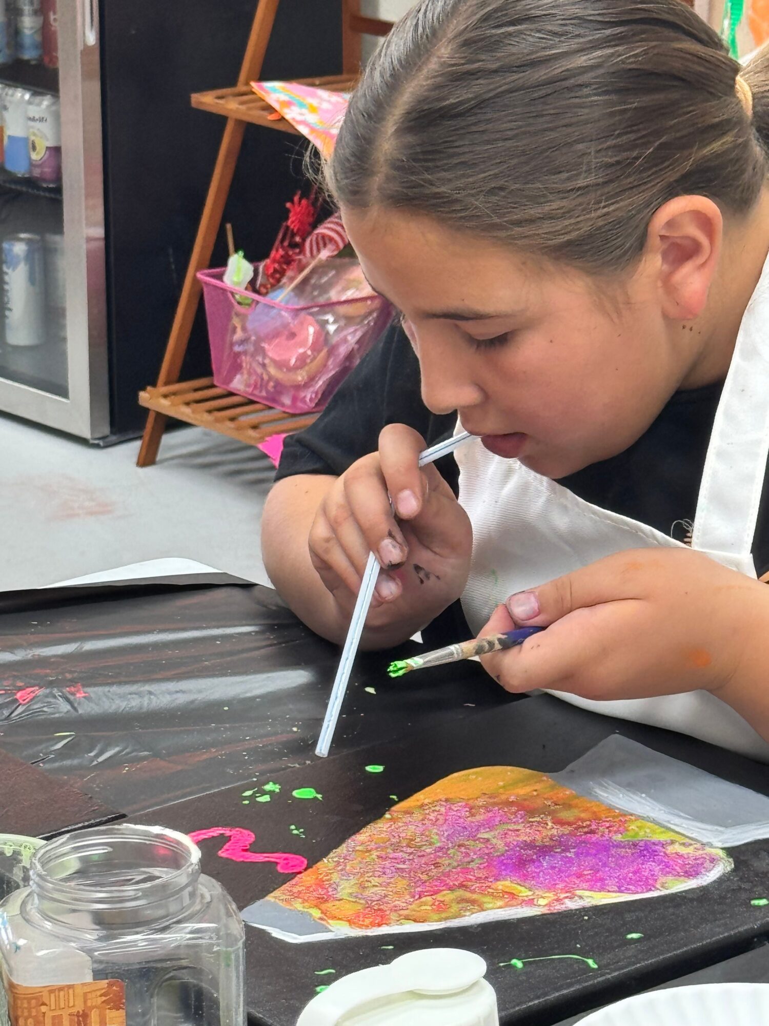Child painting a colorful heart-shaped craft with a brush, focused on the activity, on a black table with art supplies.