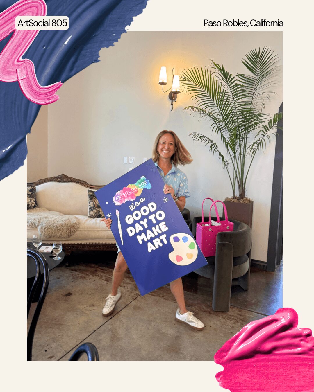 Woman holding a large blue sign with art-related text, standing in a room with a sofa, plant, and pink decorations.