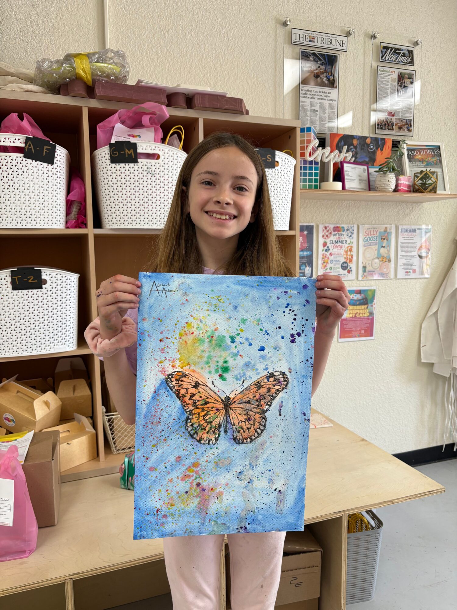 Young woman holding a colorful painting of a butterfly in a room with shelves and boxes.