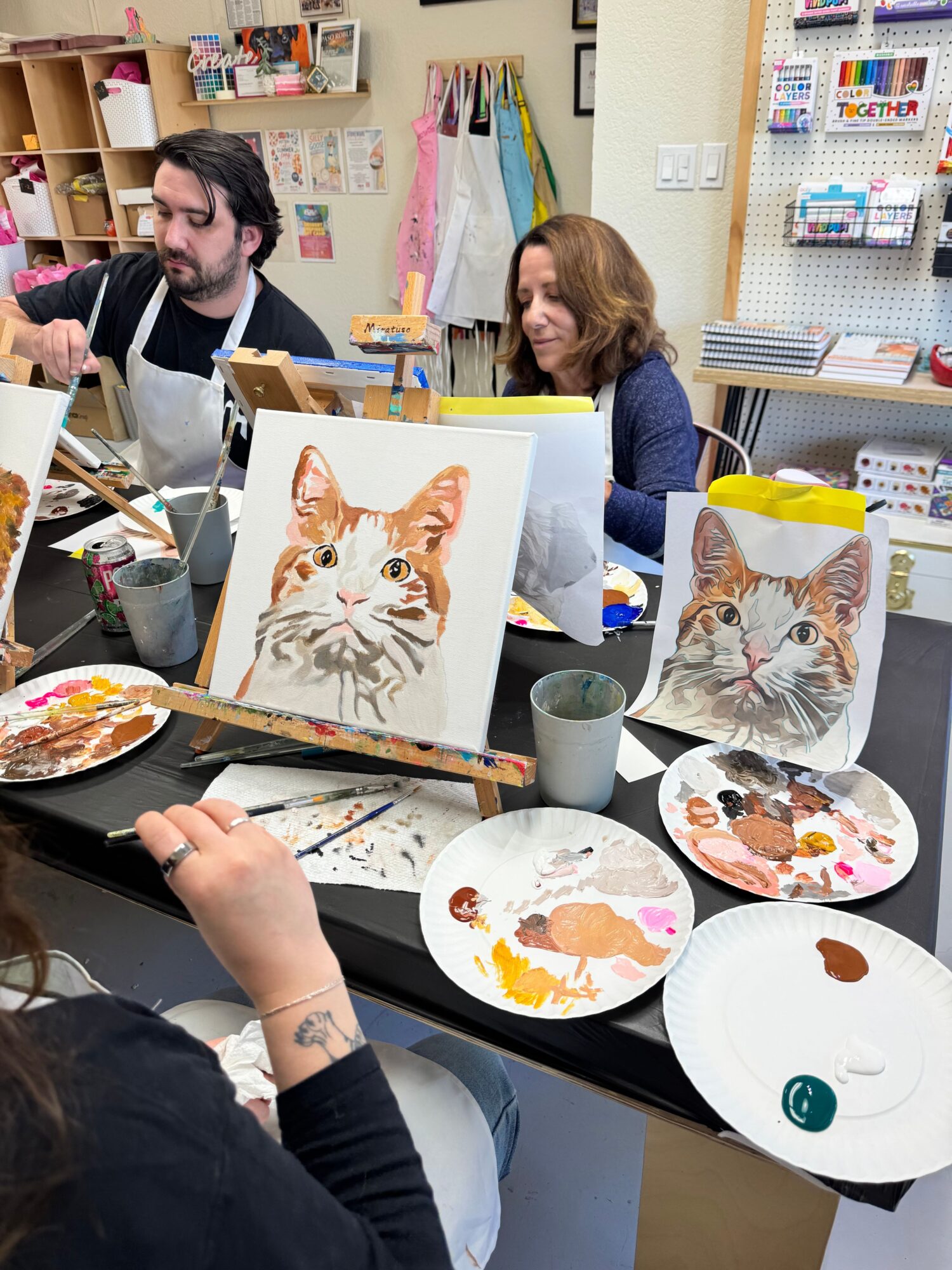 Art supplies and paintings of cats and abstract designs on a table in a classroom setting.