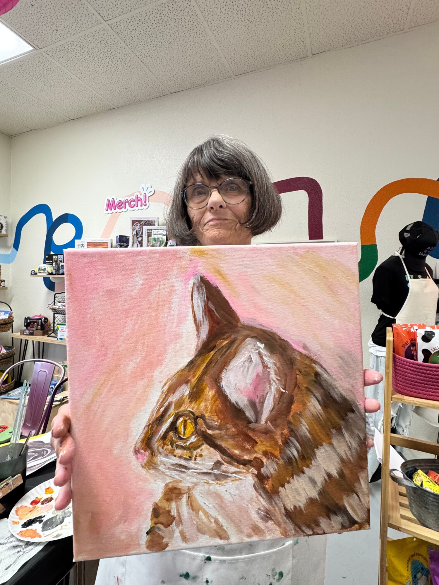 Person with gray hair holding a painting of a cow's head, with colorful background and art supplies visible.