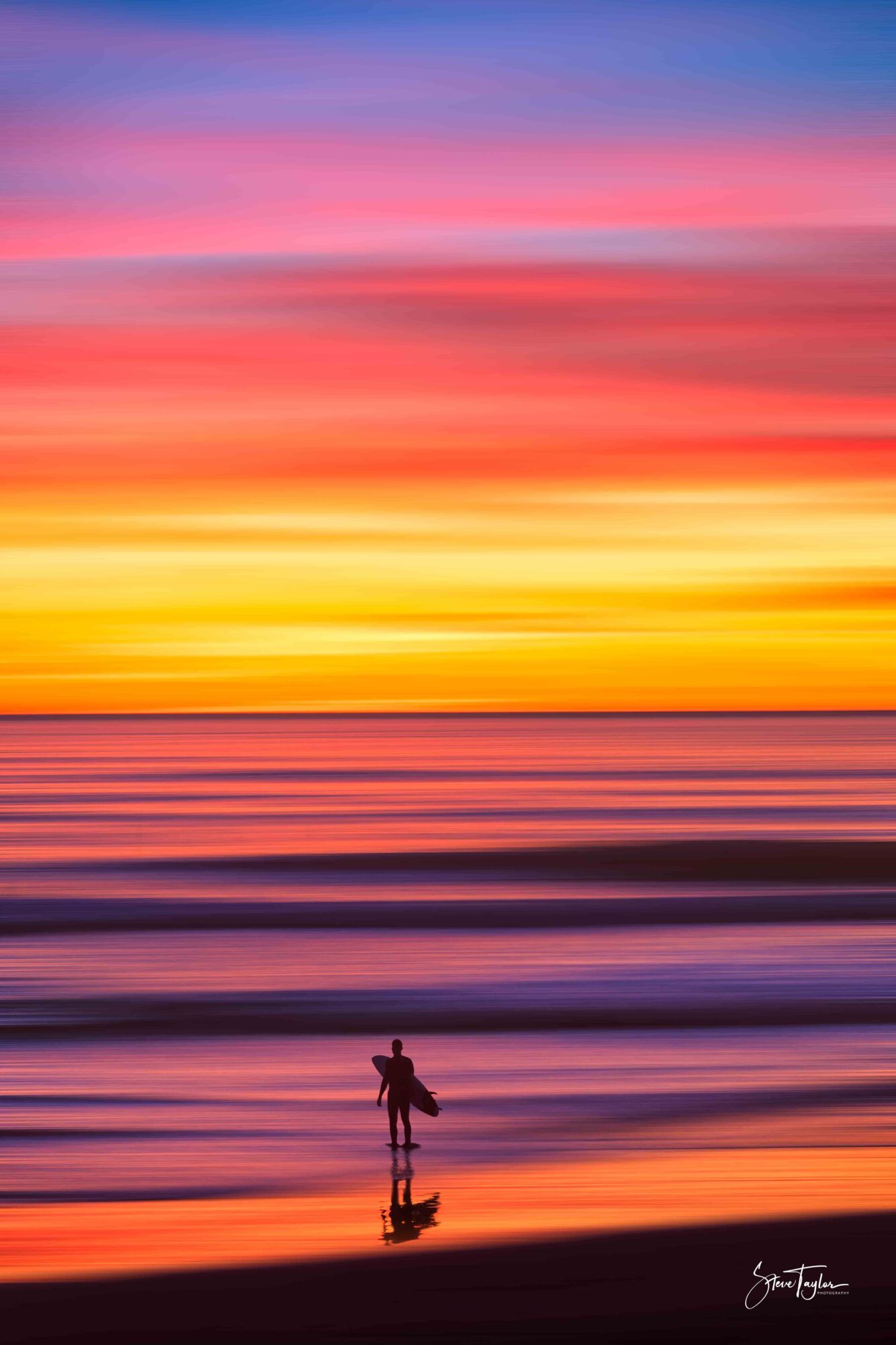 Person standing on a beach at sunset with colorful sky and reflection on water.