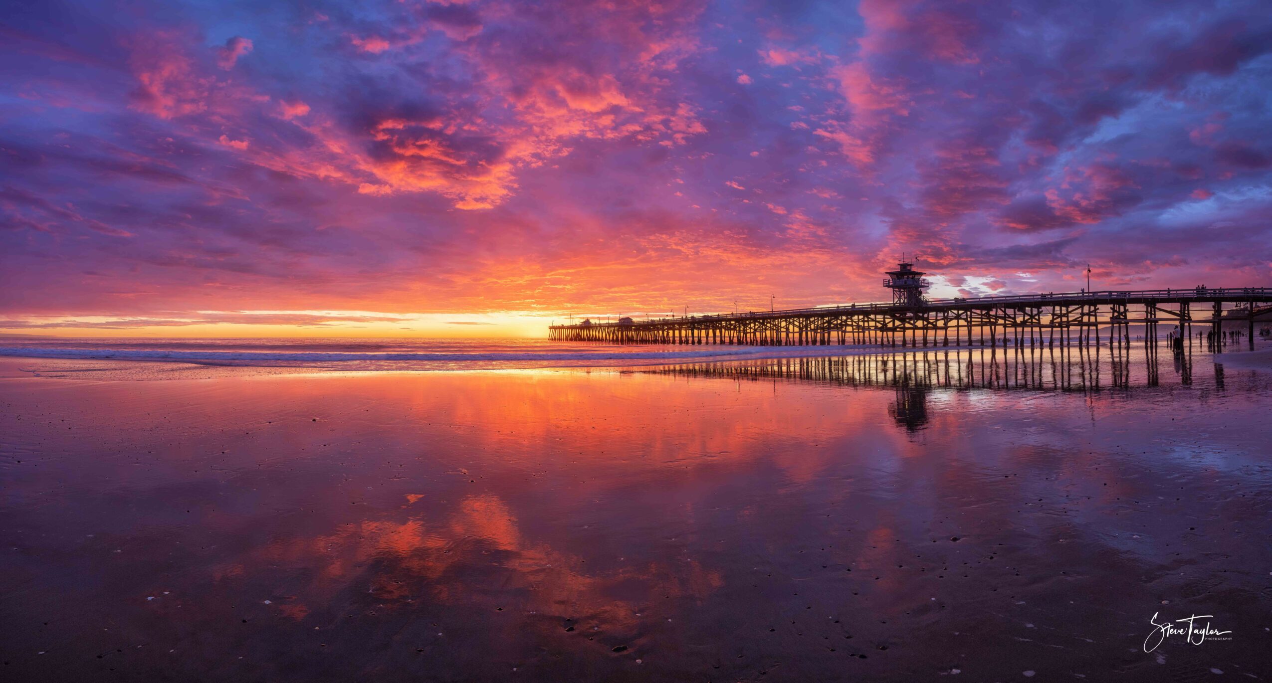 Sunset over a pier reflecting on wet sand with colorful sky and clouds, calm water, and distant horizon.