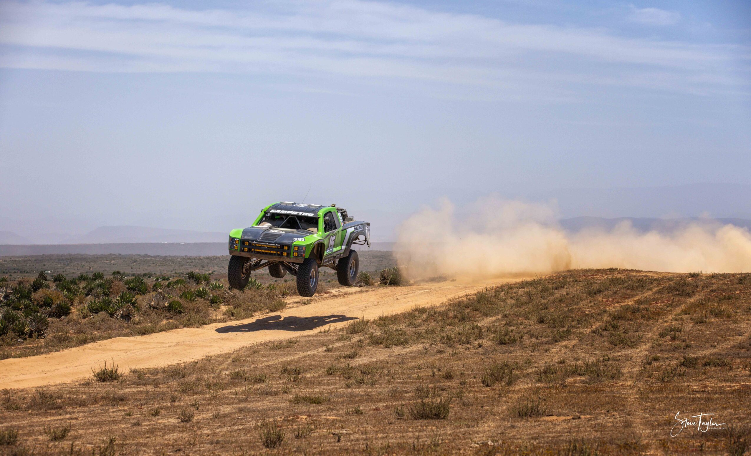 Green off-road vehicle jumps over a dirt track in a desert landscape with dust clouds behind it.