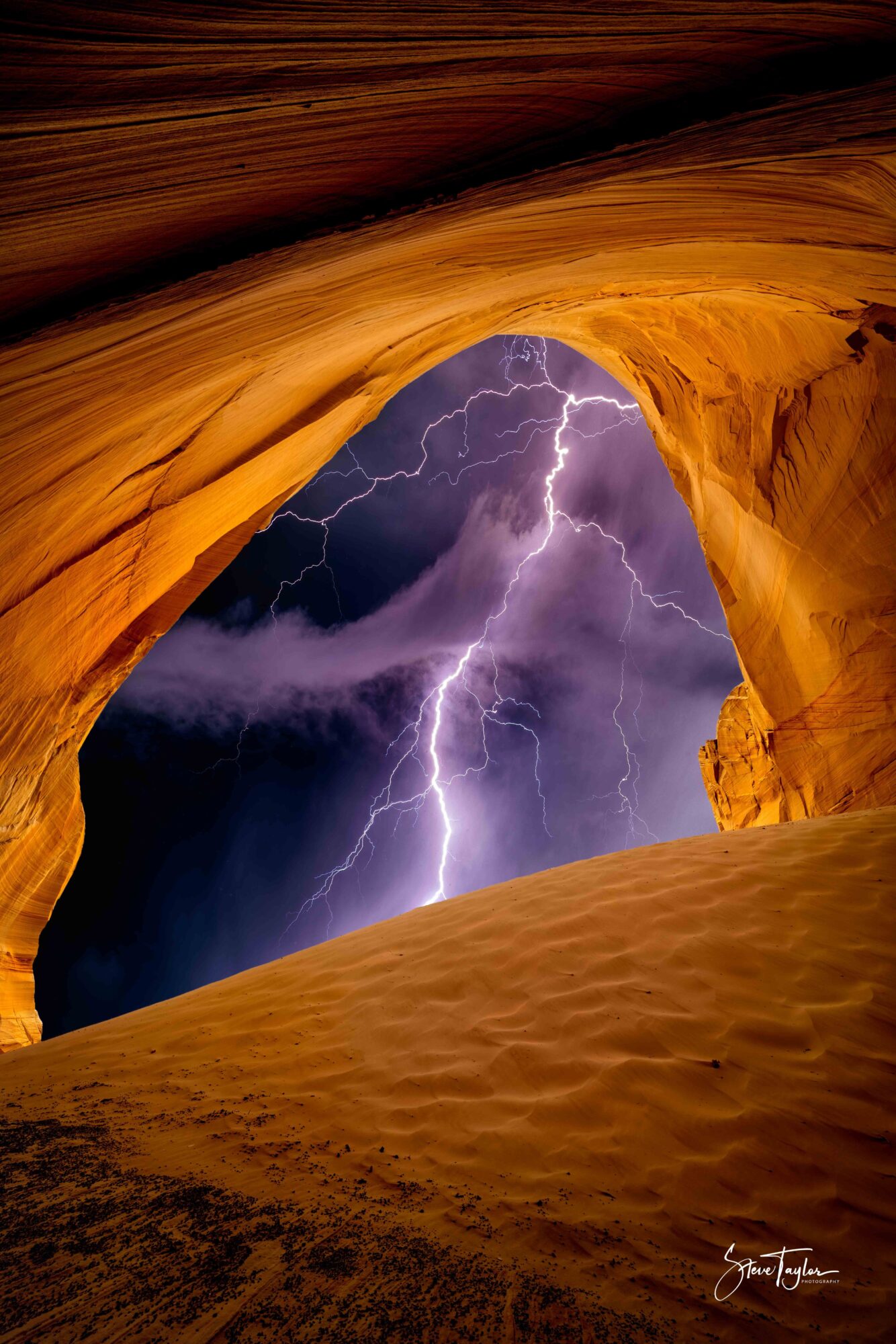 Lightning strikes through a dark stormy sky viewed from a natural rock arch formation, with sandy foreground and curved rock walls.
