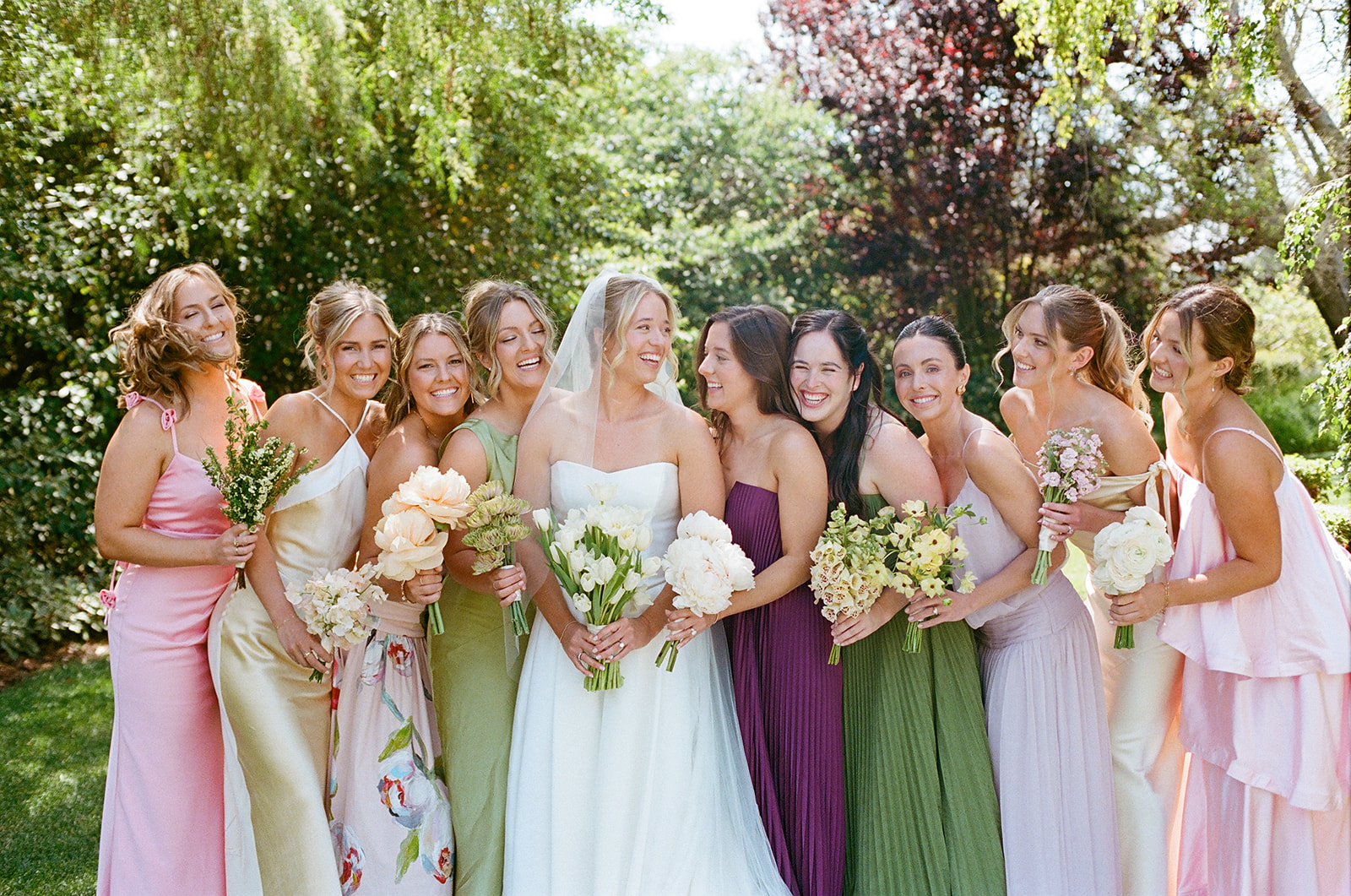 Group of women in colorful dresses outdoors, smiling, holding flowers, with trees and sunlight in background.