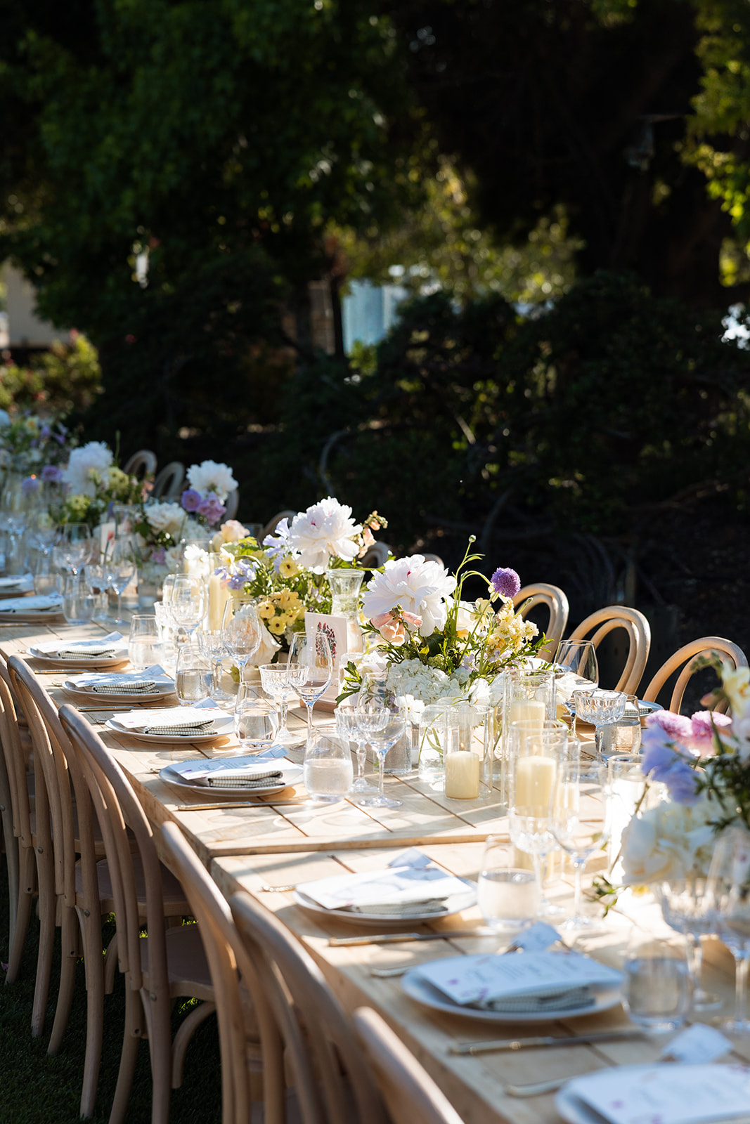 Long outdoor table set with white flowers, glassware, and napkins, surrounded by chairs, in a garden setting with trees.